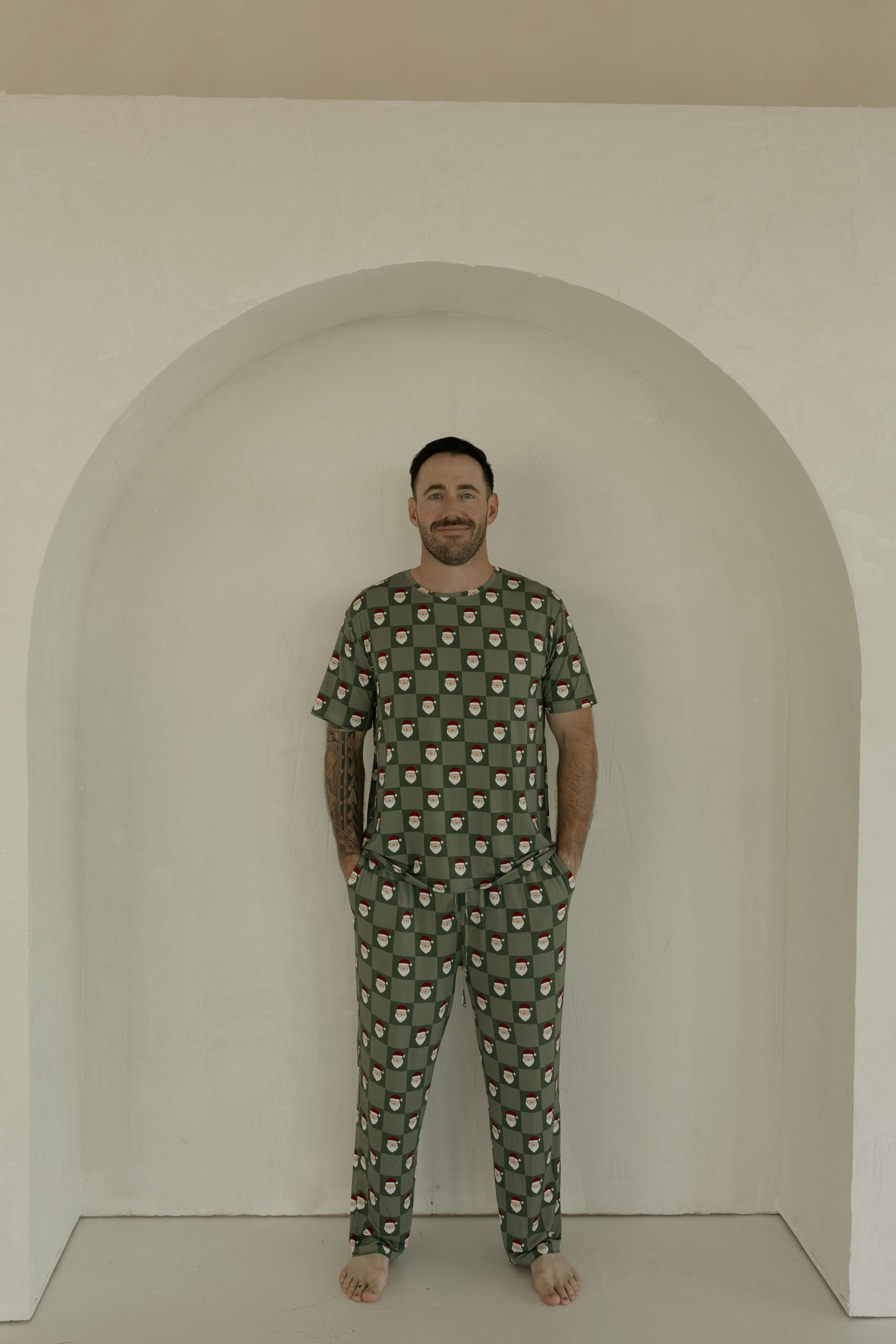 A man stands barefoot in an arched alcove, smiling at the camera with his hands in his pockets, wearing forever french baby Men's Short Sleeve Bamboo Pajamas in the Santa Checkerboard pattern.