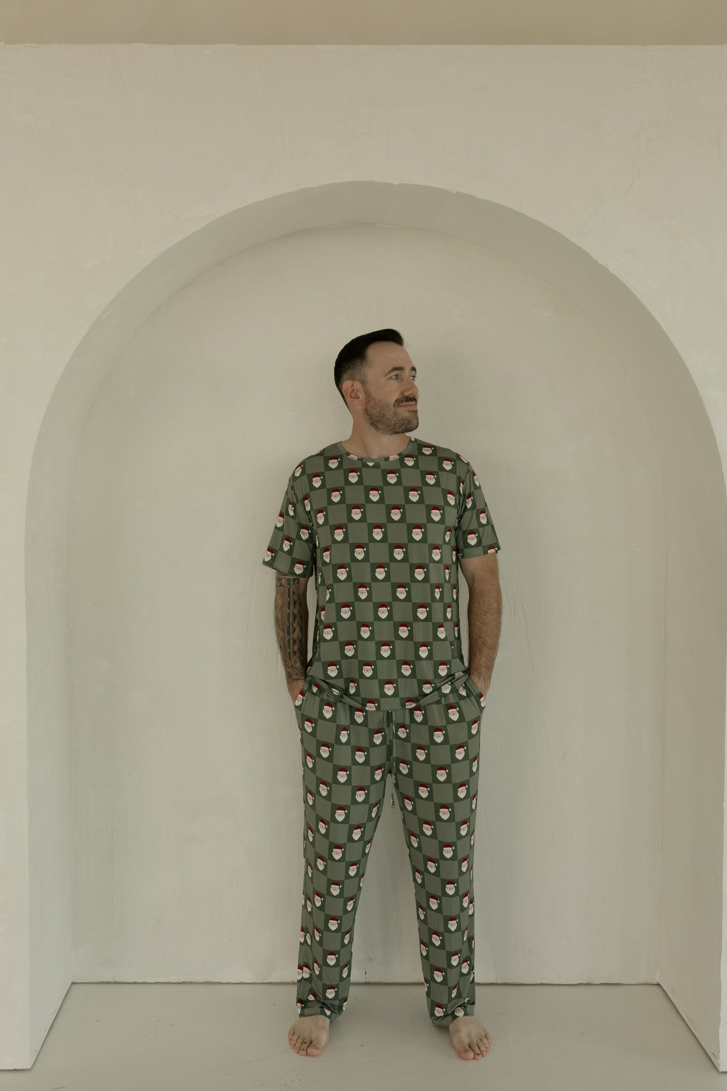 A bearded man stands barefoot in a white arched alcove, wearing forever french baby's Men's Short Sleeve Bamboo Pajamas in the Santa Checkerboard pattern. He has his hands in his pockets and looks slightly to his left.