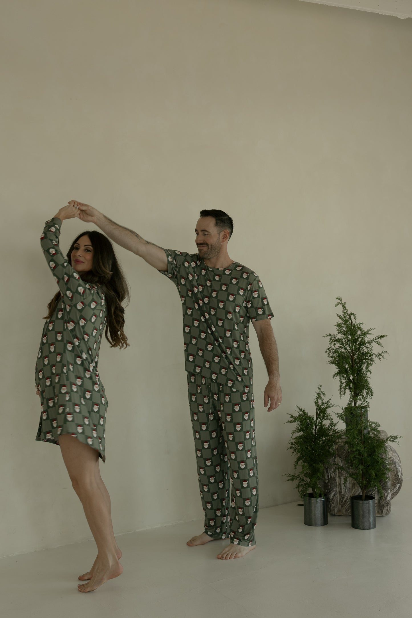 A woman in a forever french baby Women's Bamboo Sleeping Dress | Santa Checkerboard and a man, both in matching green pajamas, dance together in a minimal light room with small potted evergreen plants in the background.
