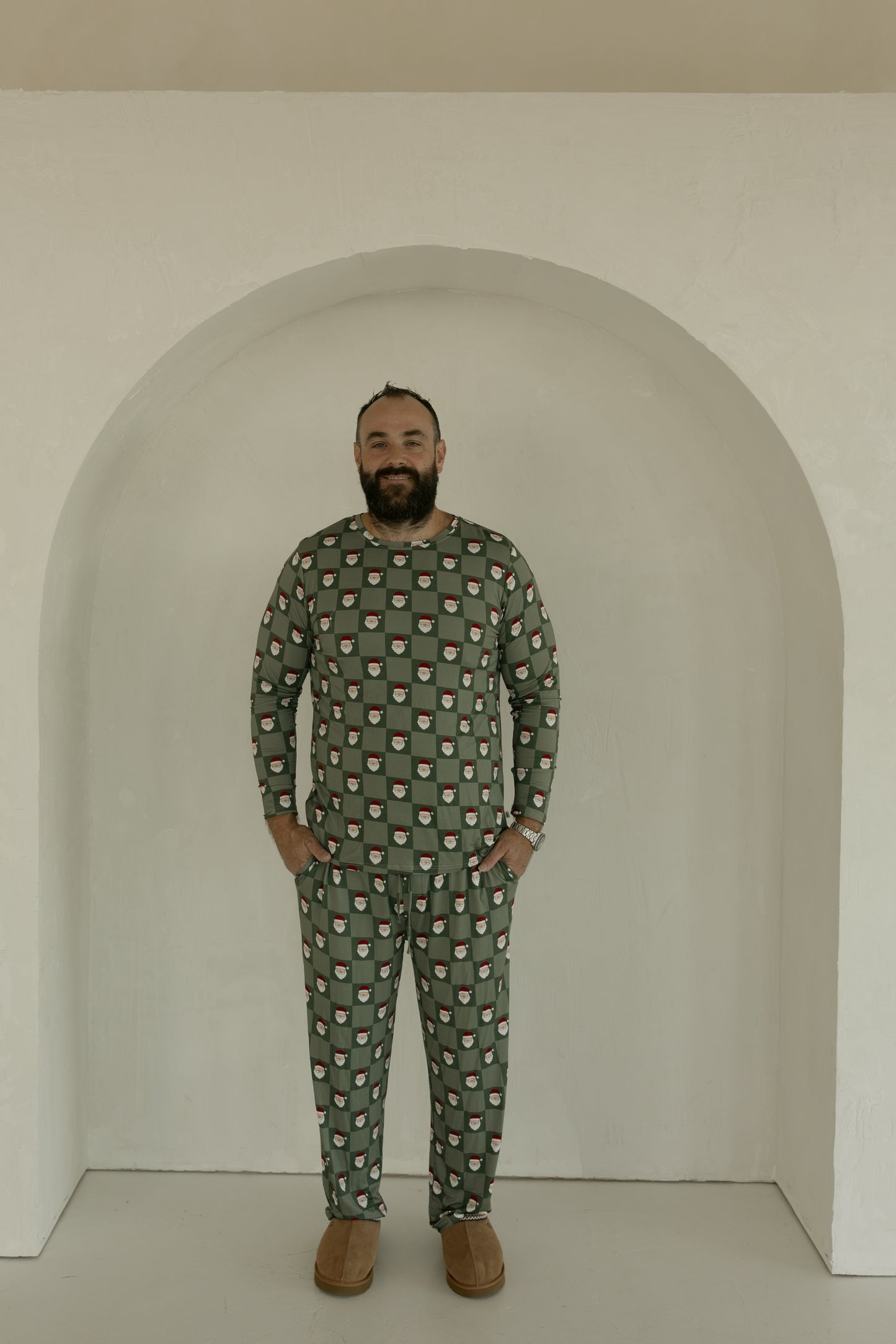 A bearded man smiles at the camera in breathable forever french baby Men's Bamboo Pajamas | Santa Checkerboard, standing before a white arched alcove and wearing green pajamas with a Santa face print and brown slippers.