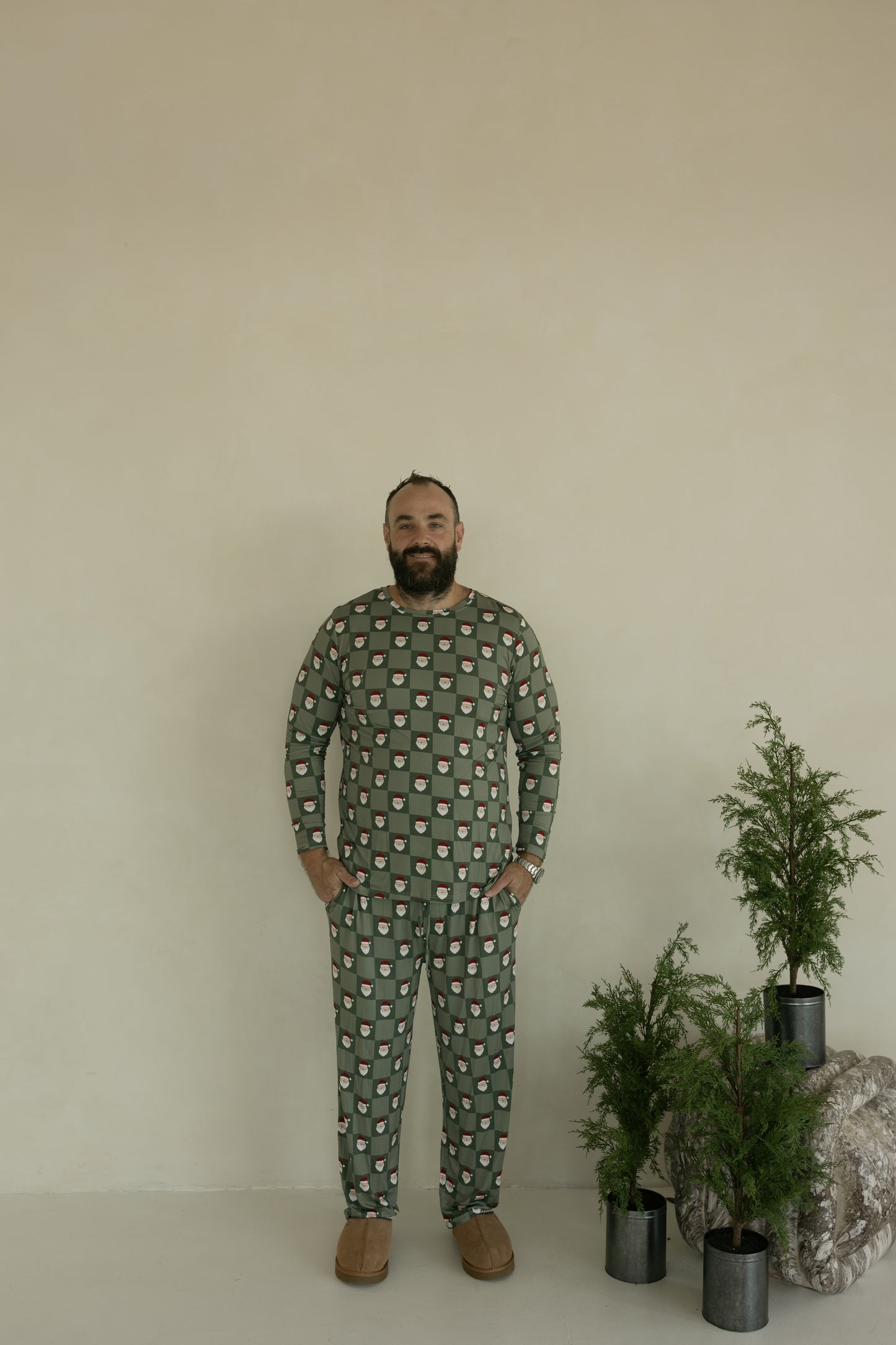 A man stands by a beige wall wearing forever french baby Men's Bamboo Pajamas in the Santa Checkerboard print and brown slippers, with small green potted plants and a rock on the floor beside him.