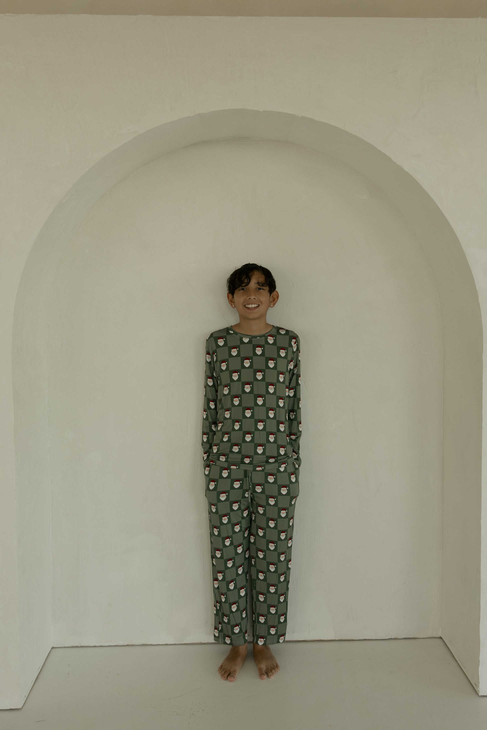 A young boy with short, dark hair stands barefoot in a white alcove, smiling in forever french baby Pre-Teen Straight Leg Bamboo Pajamas | Santa Checkerboard.