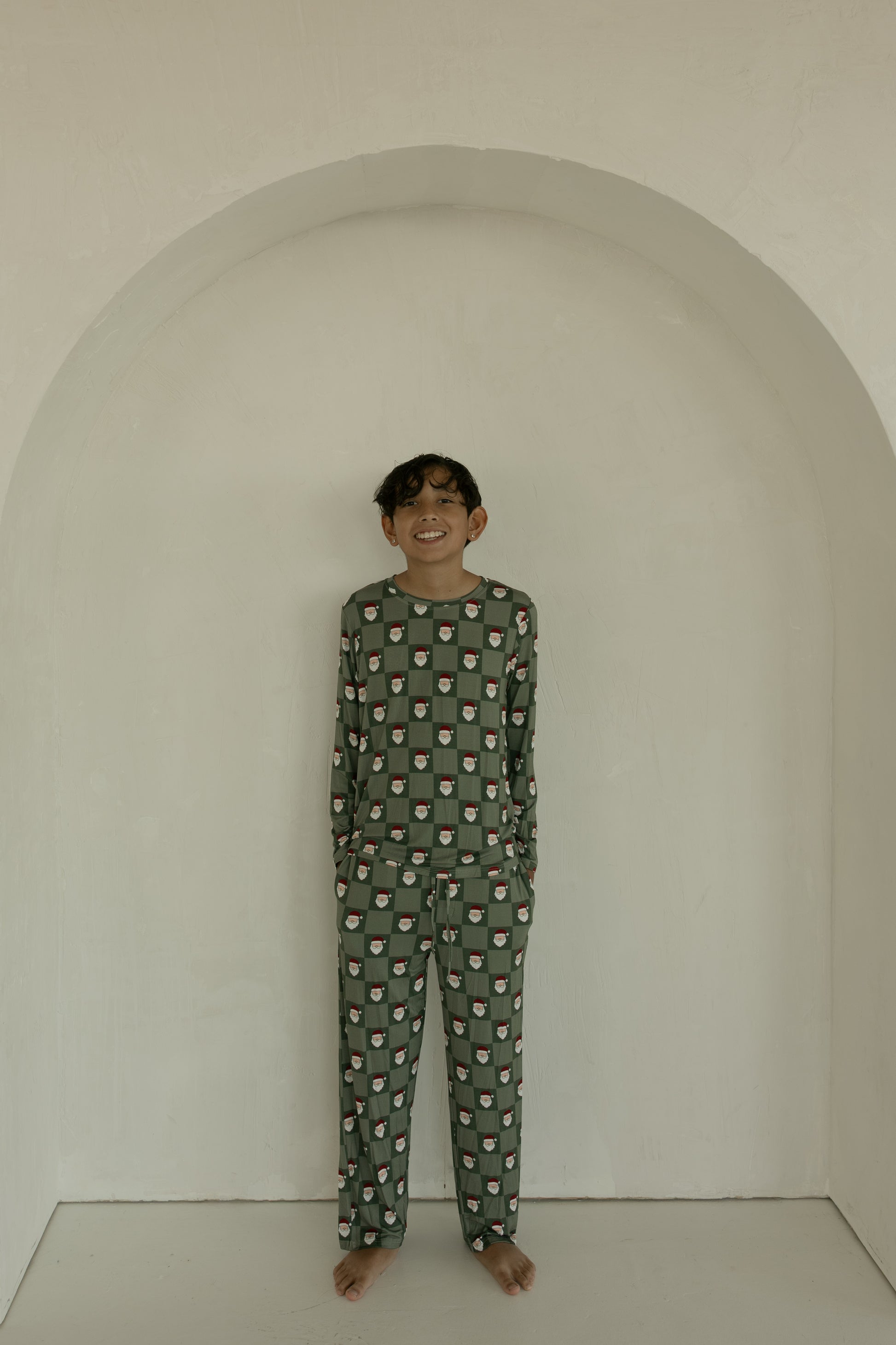 A smiling child stands barefoot by a light arched wall, wearing forever french baby’s Pre-Teen Straight Leg Bamboo Pajamas in Santa Checkerboard—a green set featuring small white and red faces.
