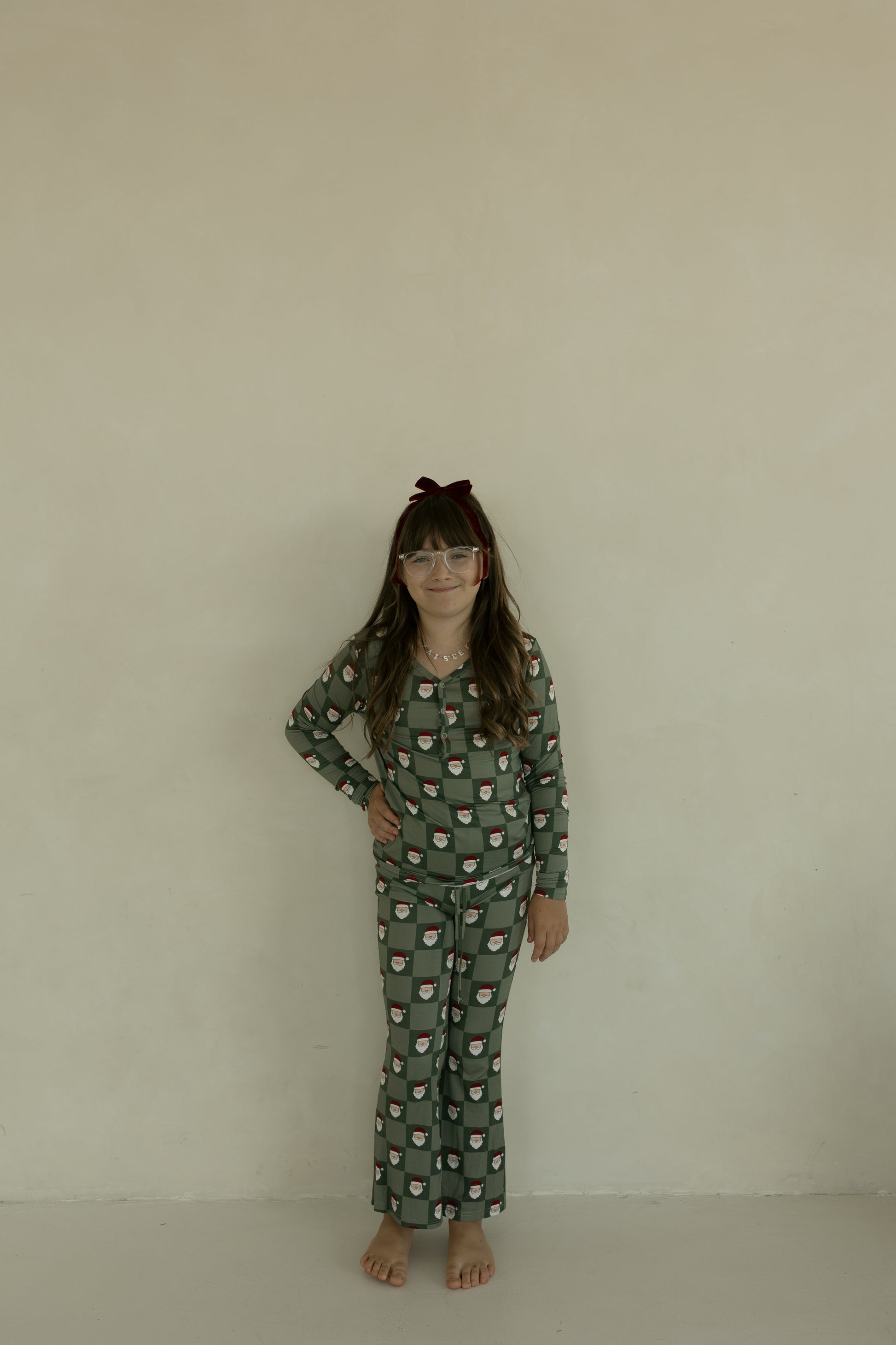A young girl in forever french baby's Pre-Teen Flare Bamboo Pajamas | Santa Checkerboard stands barefoot against a light wall, wearing glasses and a red velvet bow headband, one hand on her hip with a slight smile.