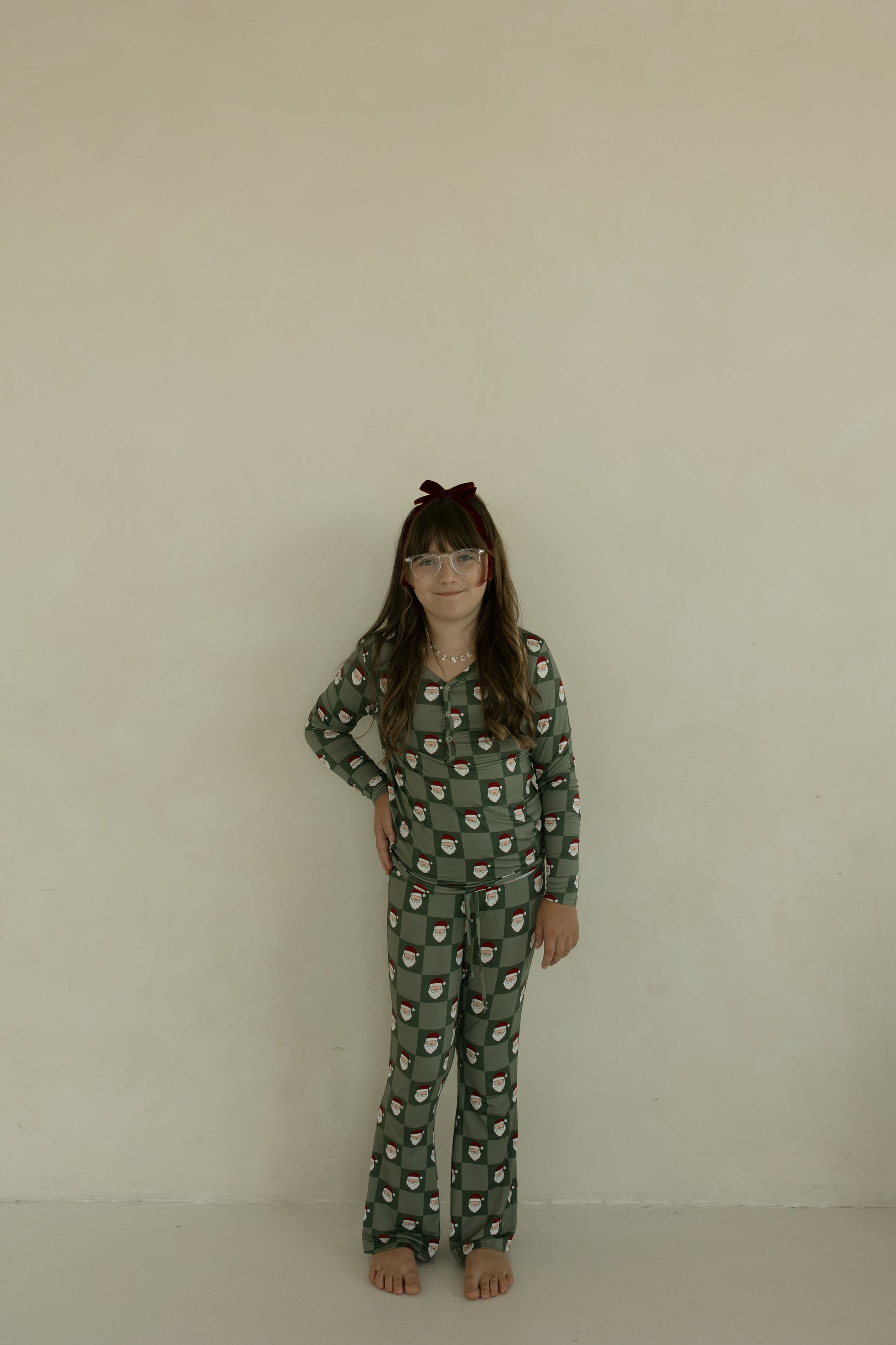A young girl with long brown hair and glasses stands barefoot against a beige wall, wearing forever french baby’s Pre-Teen Flare Bamboo Pajamas in the Santa Checkerboard print.