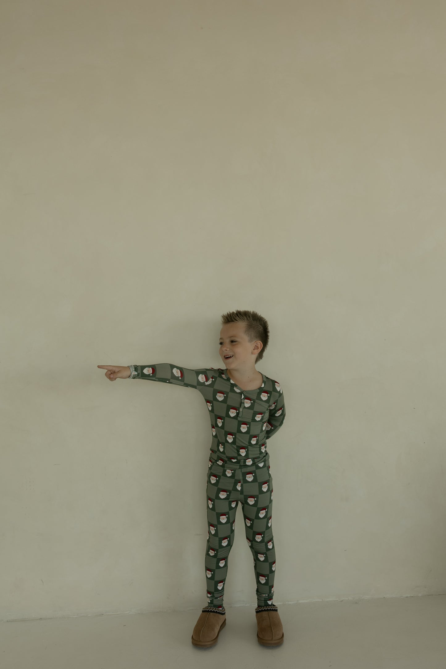 A young child smiles and points left while wearing forever french baby's Bamboo Two Piece Pajamas | Santa Checkerboard, standing against a beige wall. The breathable pajamas are paired with light brown slippers for added comfort.