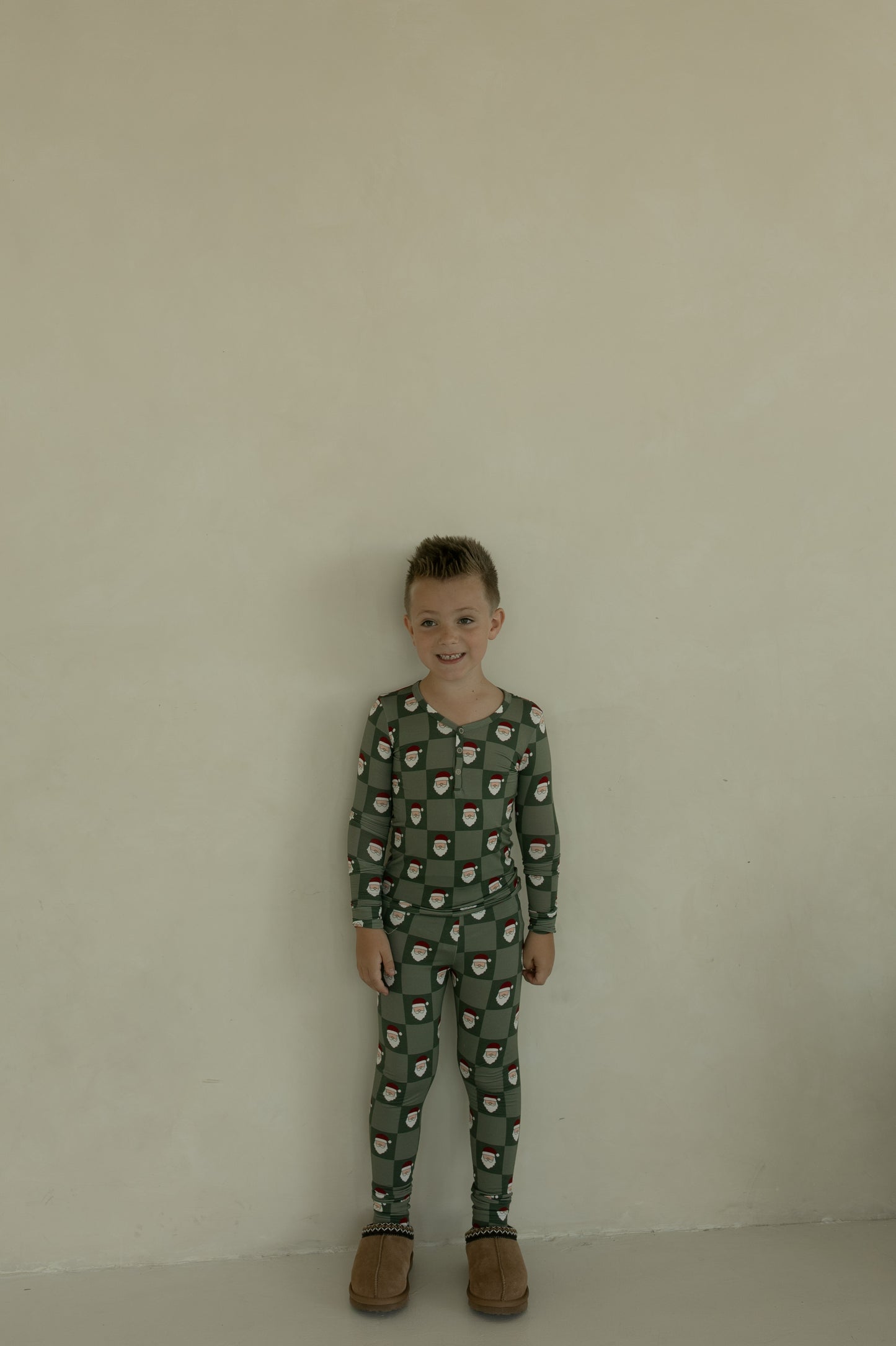 A young boy stands against a plain light-colored wall, smiling at the camera in forever french baby’s Bamboo Two Piece Pajamas | Santa Checkerboard and oversized brown slippers.