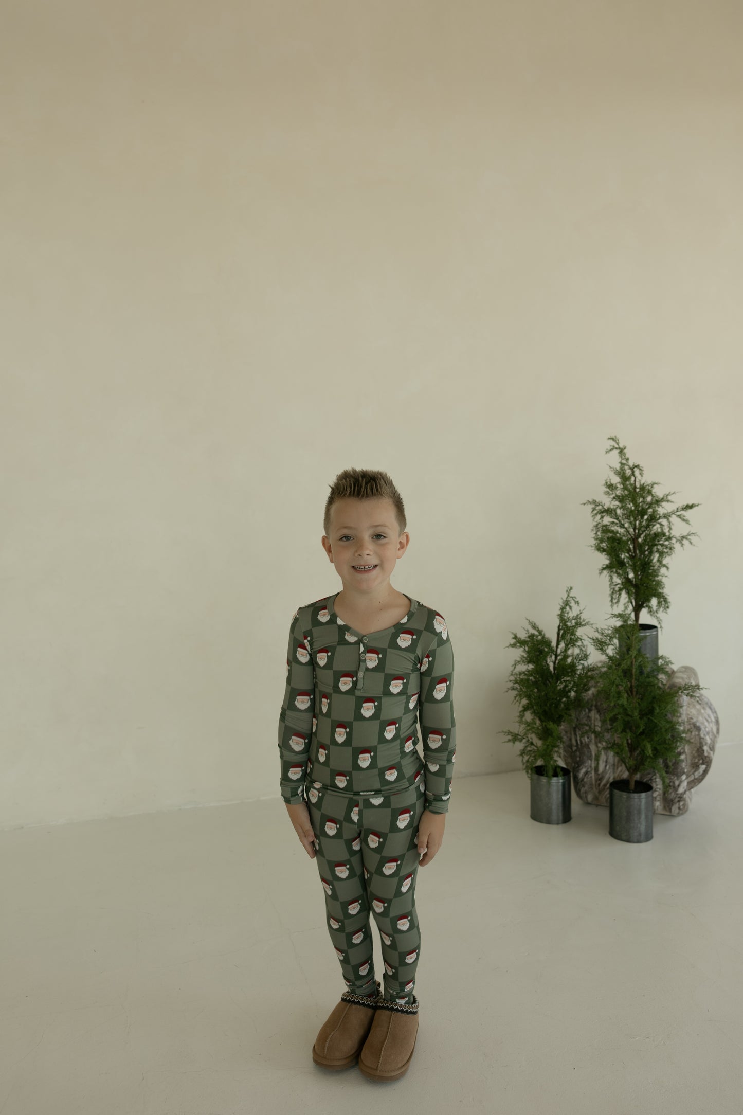 A young child wears forever french baby’s Bamboo Two Piece Pajamas in Santa Checkerboard, standing indoors on a light floor with small potted evergreens on rocks and a plain beige wall in the background.