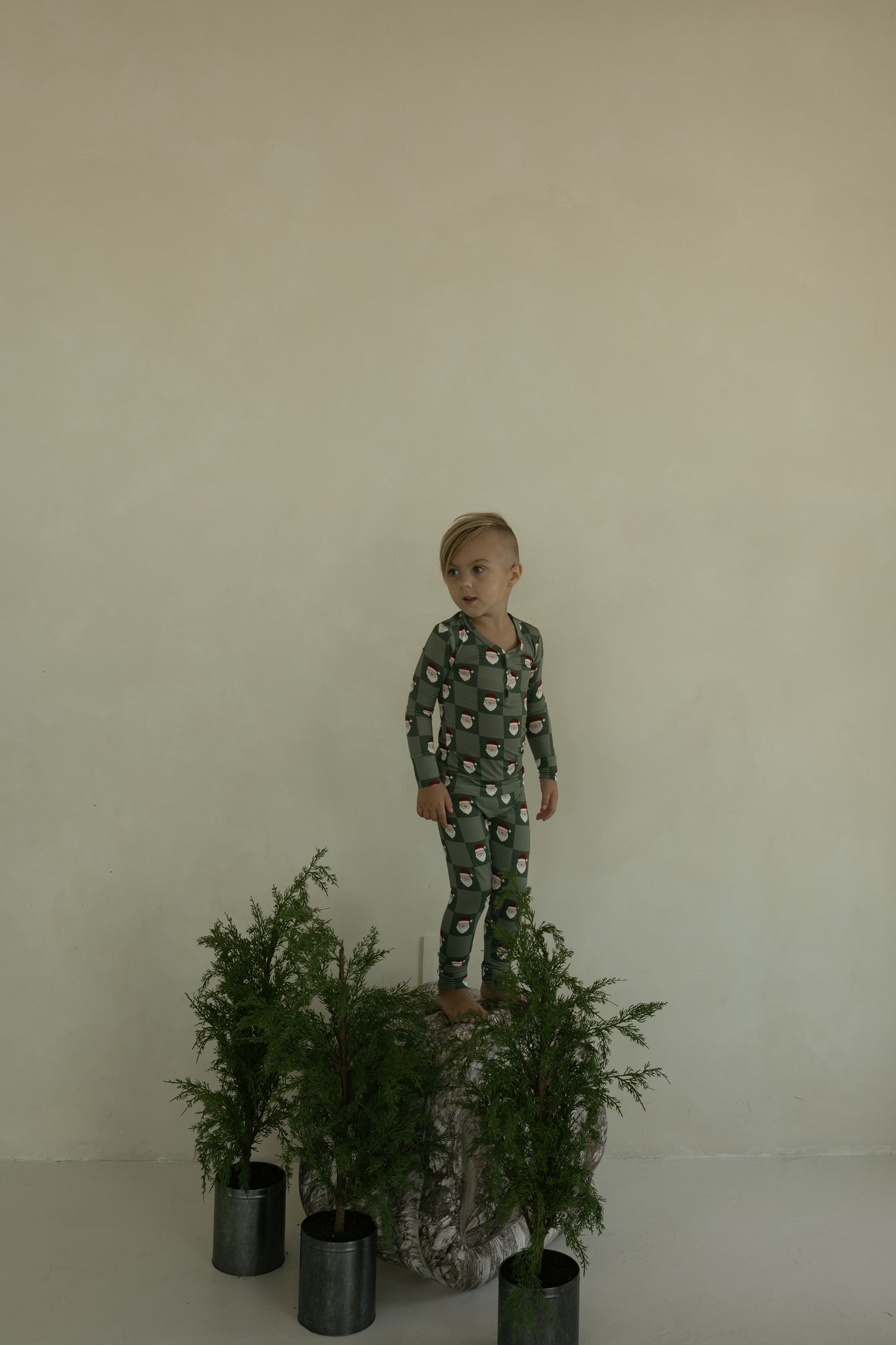 A young child in forever french baby's hypo-allergenic Bamboo Two Piece Pajamas | Santa Checkerboard stands barefoot on a tree stump among potted plants, wearing green patterned pajamas against a plain beige wall.