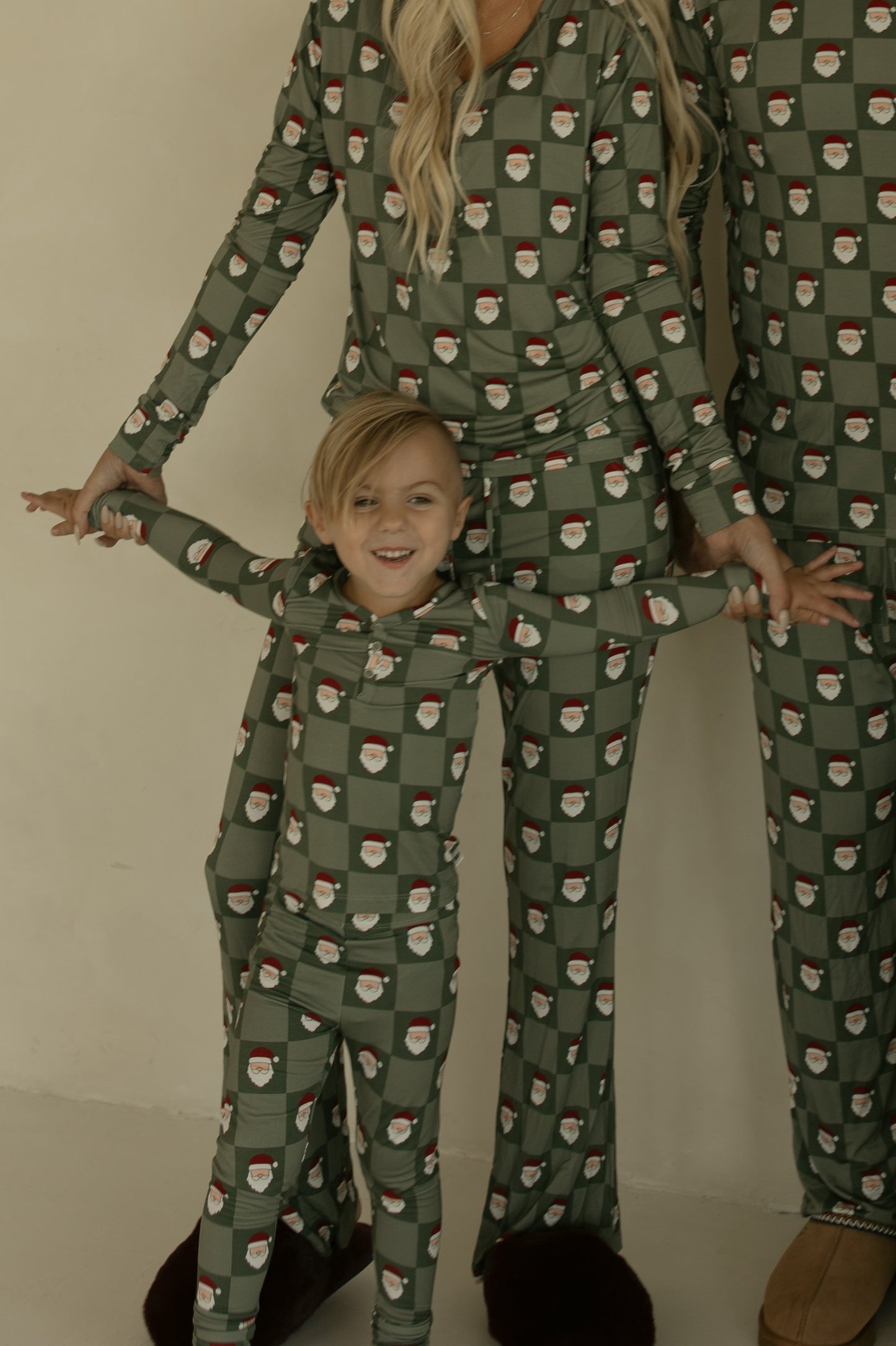 A smiling child and two adults wear matching forever french baby Bamboo Two Piece Pajamas | Santa Checkerboard—soft, breathable sleepwear in green with a Santa pattern—standing together by a light wall, with adults' faces partly out of frame.