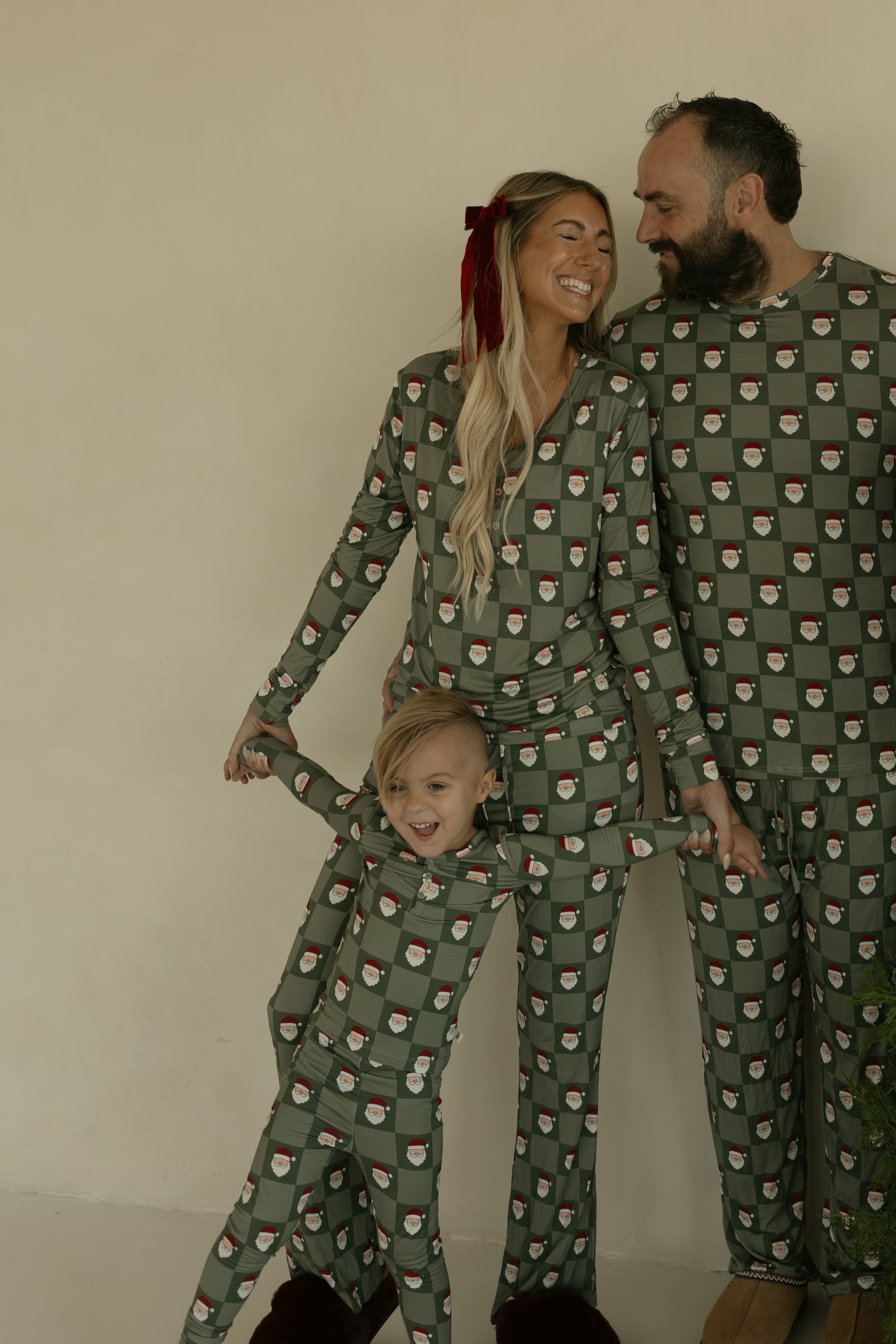 A smiling family of three wears forever french baby Bamboo Two Piece Pajamas | Santa Checkerboard—matching green, breathable sleepwear with a festive print as their child stretches out, grinning.