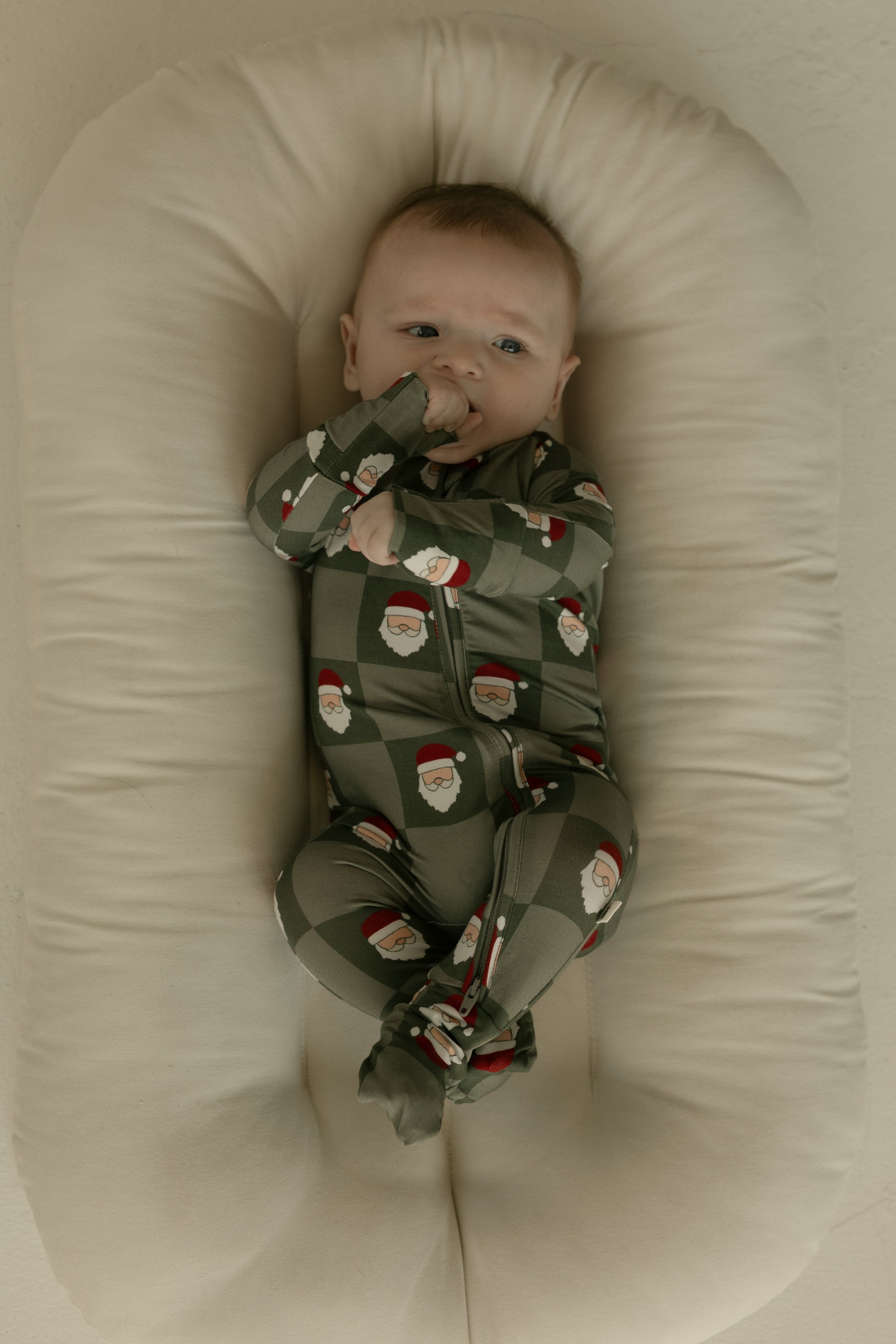 A baby in forever french baby's Bamboo Zip Pajamas | Santa Checkerboard relaxes on a soft off-white cushion, sucking their thumb and gazing upward.