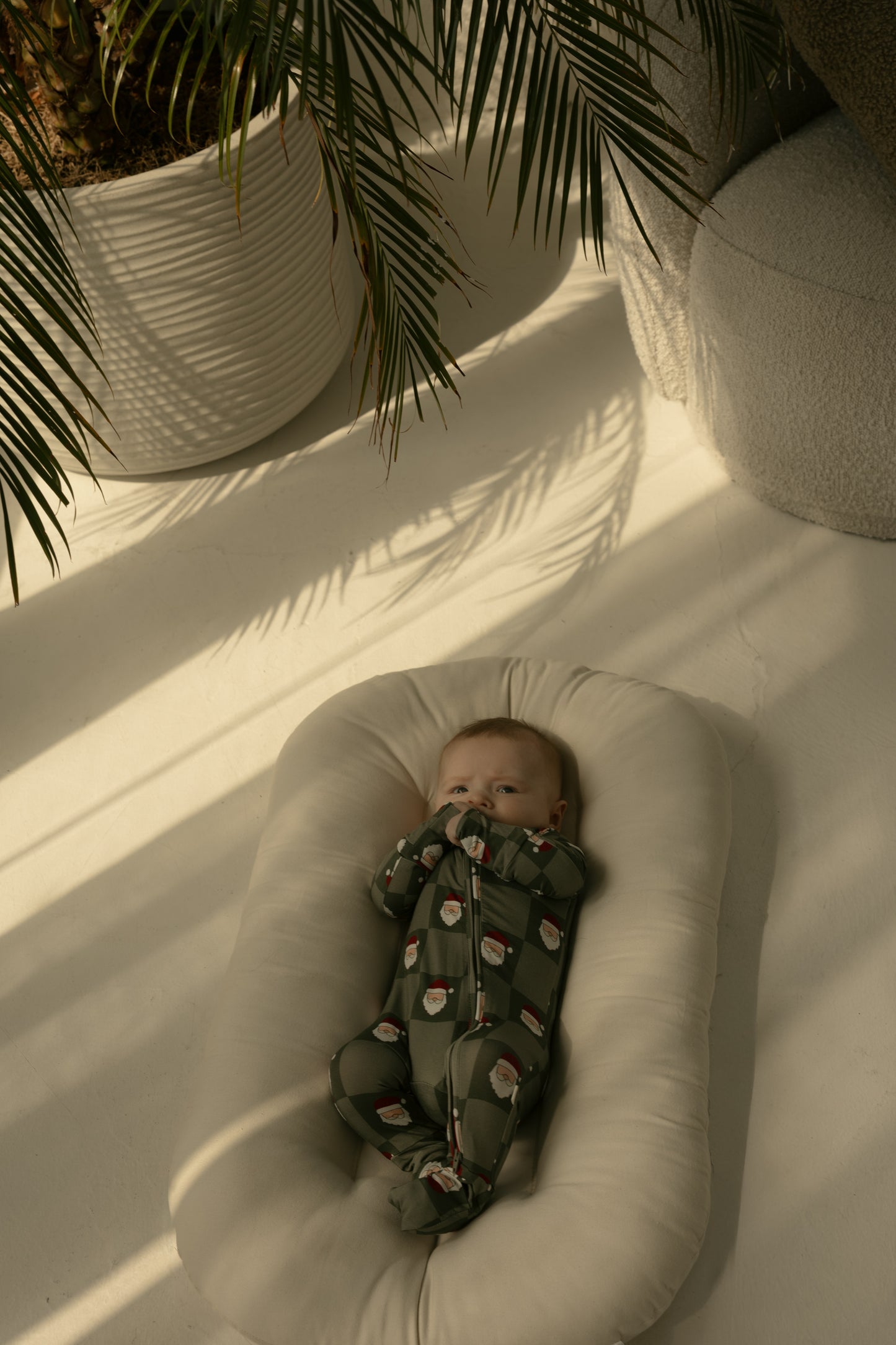A baby wearing forever french baby Bamboo Zip Pajamas | Santa Checkerboard lies on a soft white cushion in sunlight. Nearby, large potted plants cast gentle shadows, creating a cozy, peaceful scene.