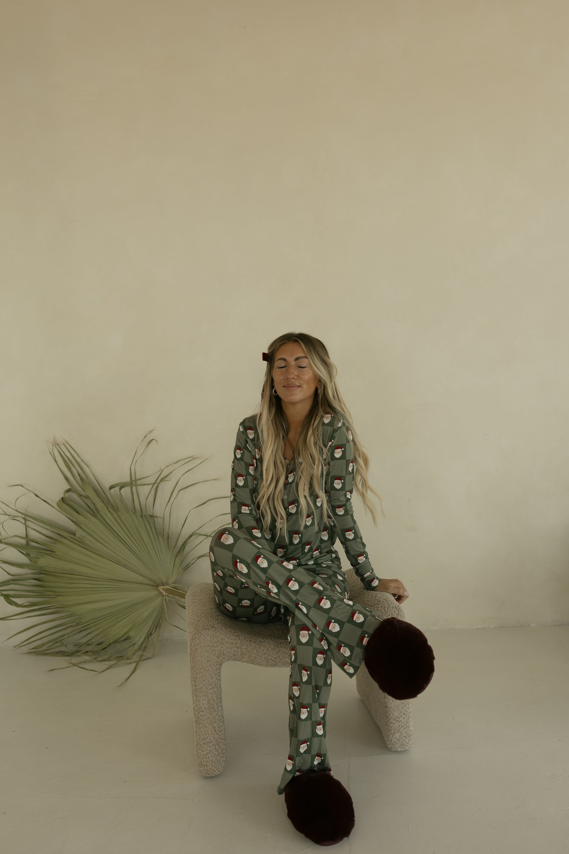 A woman with long blonde hair, eyes closed, relaxes on a textured beige chair in forever french baby's Women's Bamboo Pajamas | Santa Checkerboard. Large dark slippers cover her feet, and a dried palm leaf rests nearby.