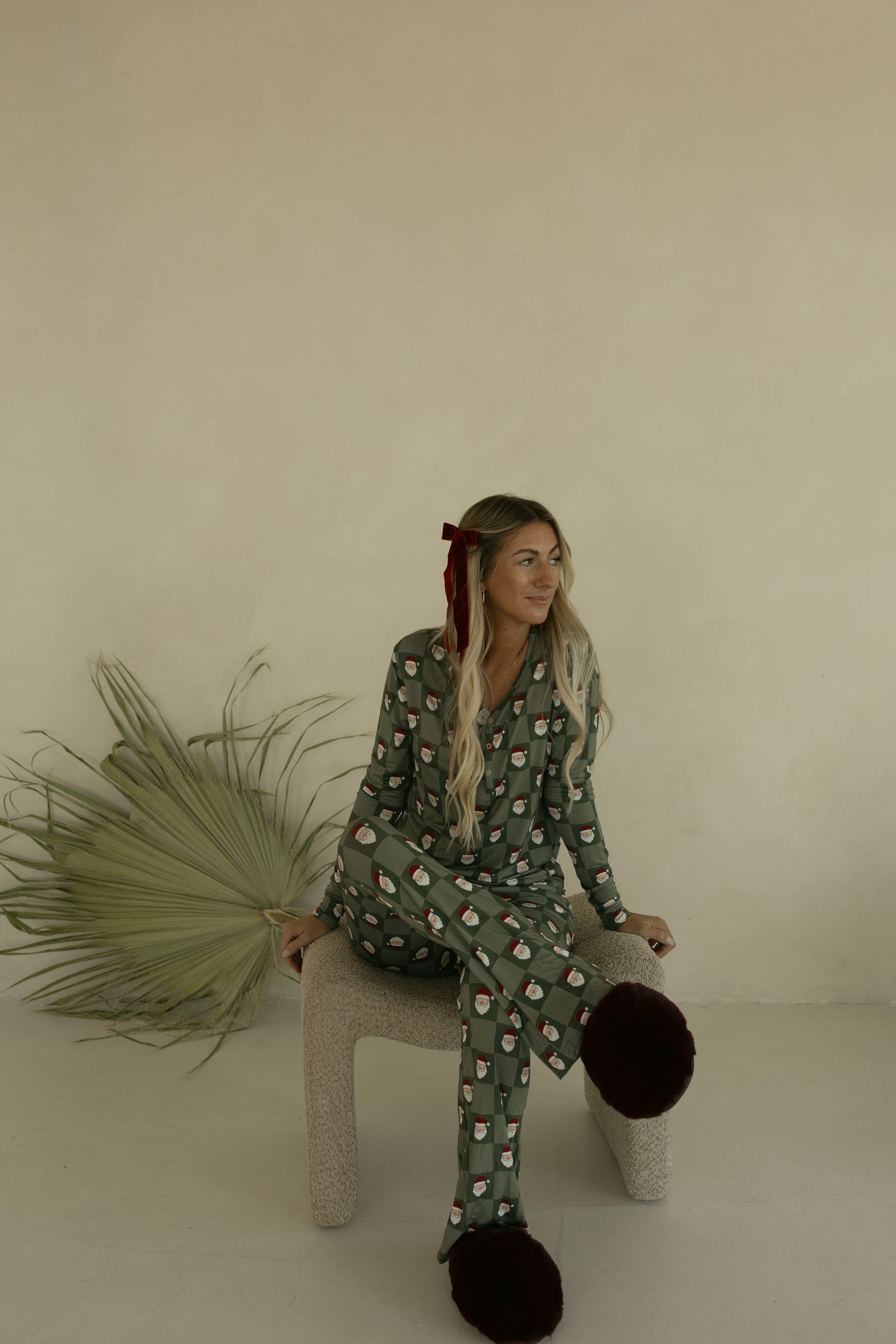 A woman with long blonde hair, wearing forever french baby Women's Bamboo Pajamas | Santa Checkerboard and large brown slippers, sits on a textured chair. A dried palm leaf rests against the beige wall behind her.