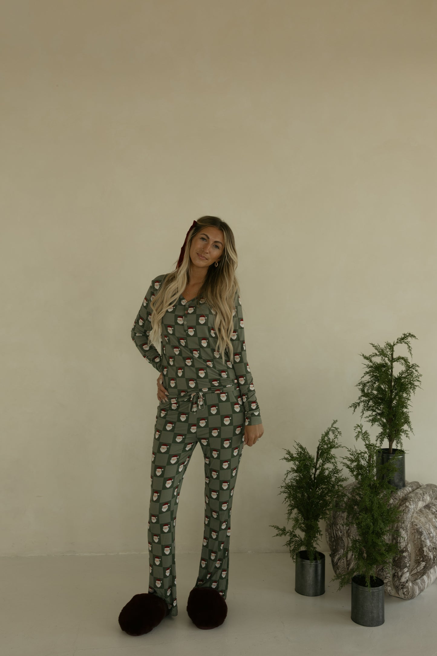 A woman with long blonde hair stands indoors in forever french baby Women's Bamboo Pajamas | Santa Checkerboard, paired with large dark slippers. She is surrounded by small potted evergreens against a beige wall.