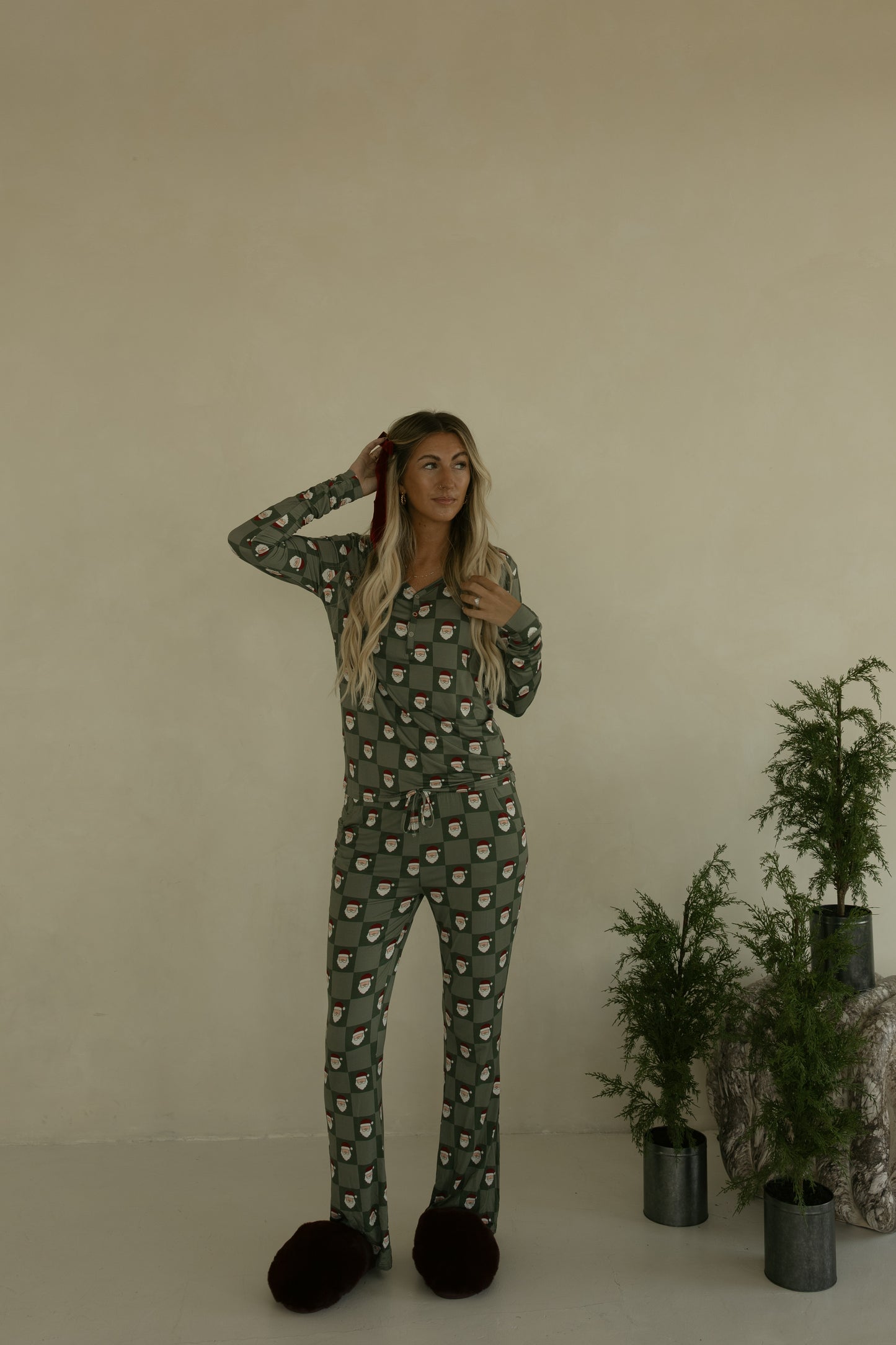 A woman with long blonde hair stands indoors in forever french baby Women's Bamboo Pajamas | Santa Checkerboard, wearing dark slippers. She touches her hair near green plants against a beige wall, enjoying the hypoallergenic comfort.