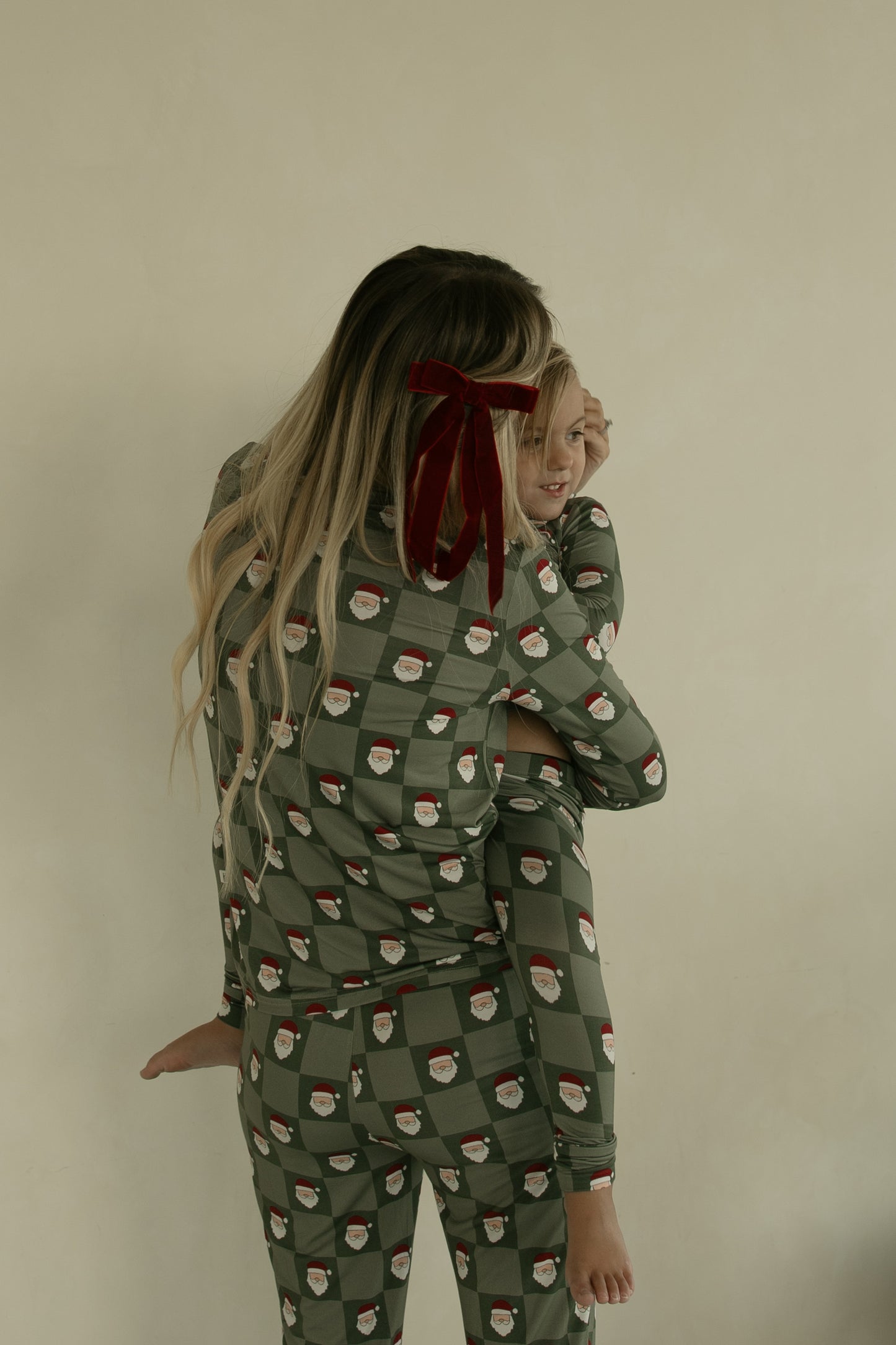 A woman with long blonde hair tied with a red velvet bow holds a smiling child. Both wear forever french baby Women's Bamboo Pajamas in the Santa Checkerboard pattern, standing against a light wall.
