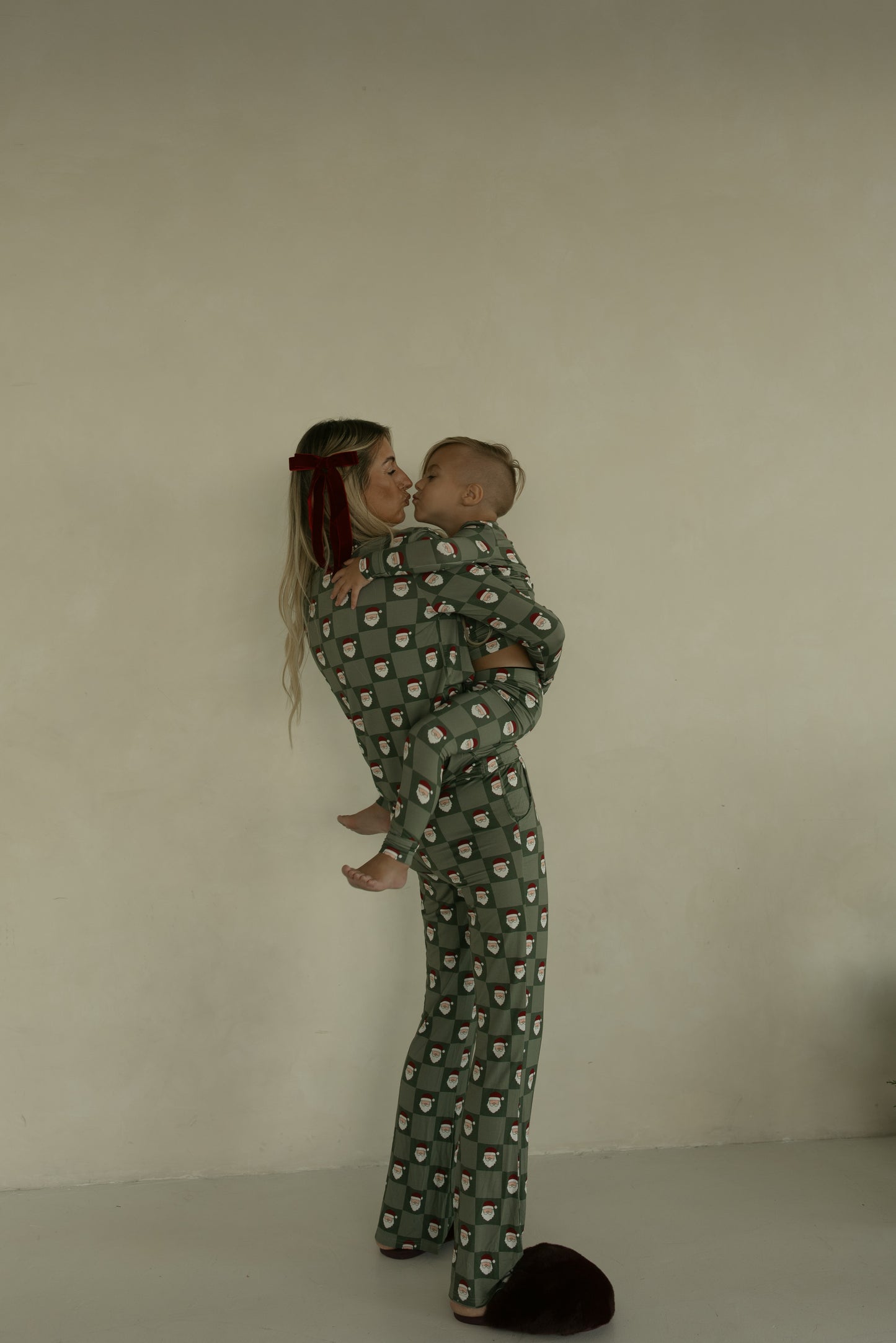 A woman in forever french baby Women's Bamboo Pajamas | Santa Checkerboard, which are breastfeeding friendly, holds a small child in matching festive green sleepwear against a beige wall. She smiles as the child looks at her.
