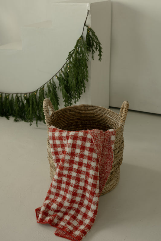 A woven basket with handles holds the forever French baby Plush Blanket | Red Gingham. In the background, a green garland with small lights decorates white stairs and a wall.