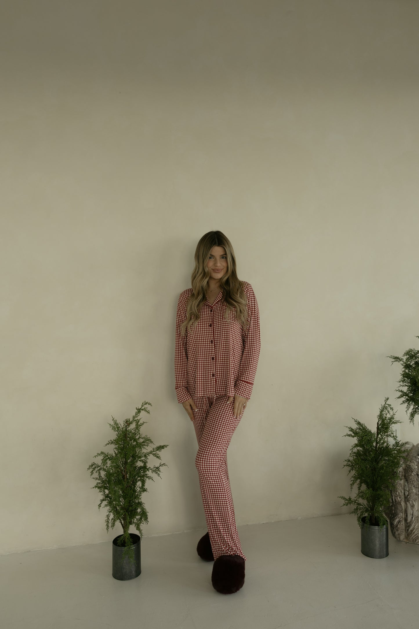 A woman with long wavy hair wears the forever french baby Long Sleeve Women's Bamboo Pajamas in Red Gingham and dark slippers, standing indoors against a beige wall with two small potted green plants.