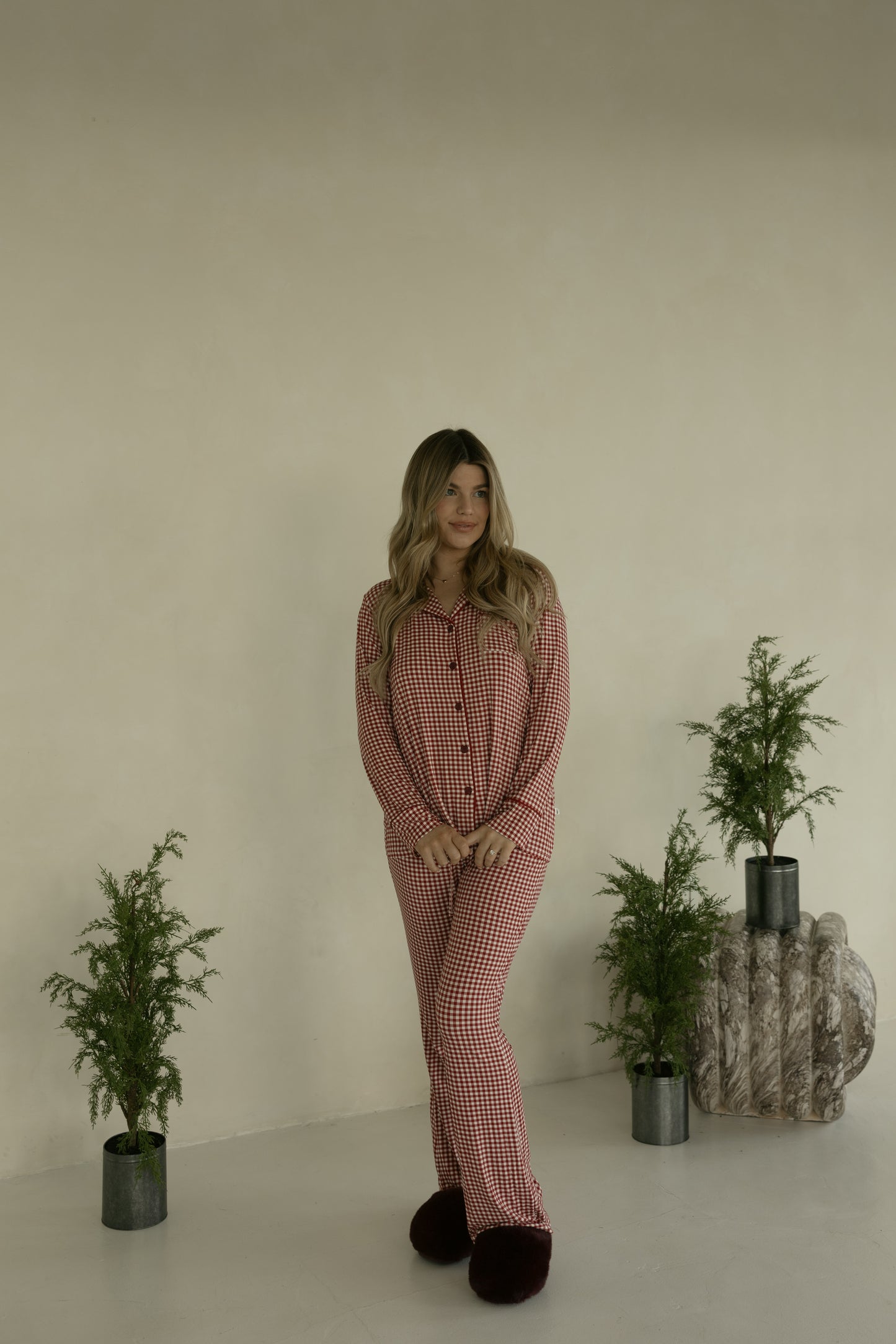 A woman with long hair stands indoors wearing forever french baby’s Long Sleeve Women's Bamboo Pajamas in Red Gingham and dark slippers, surrounded by three small potted plants against a light-colored wall.