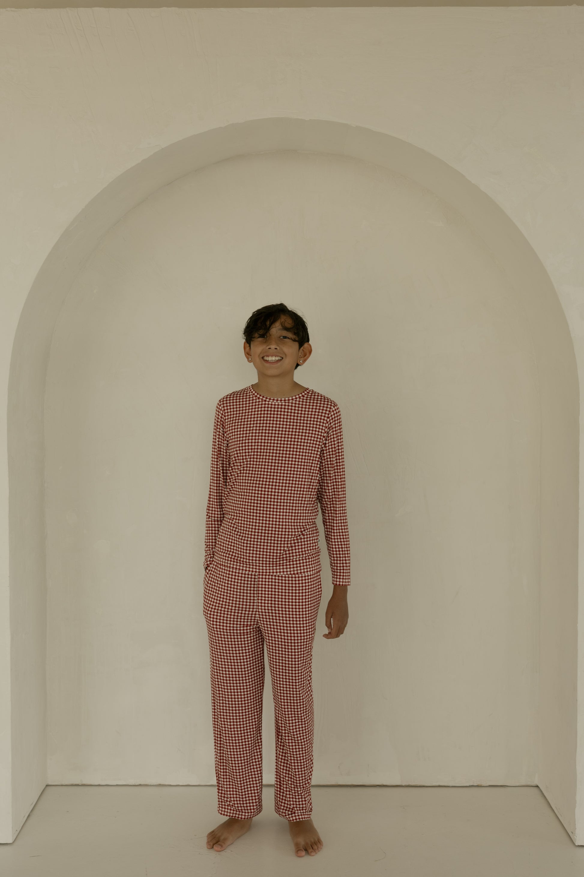 A smiling child stands barefoot under a white arch, wearing forever french baby's Pre-Teen Straight Leg Bamboo Pajamas in Red Gingham. The background is minimal and neutral.