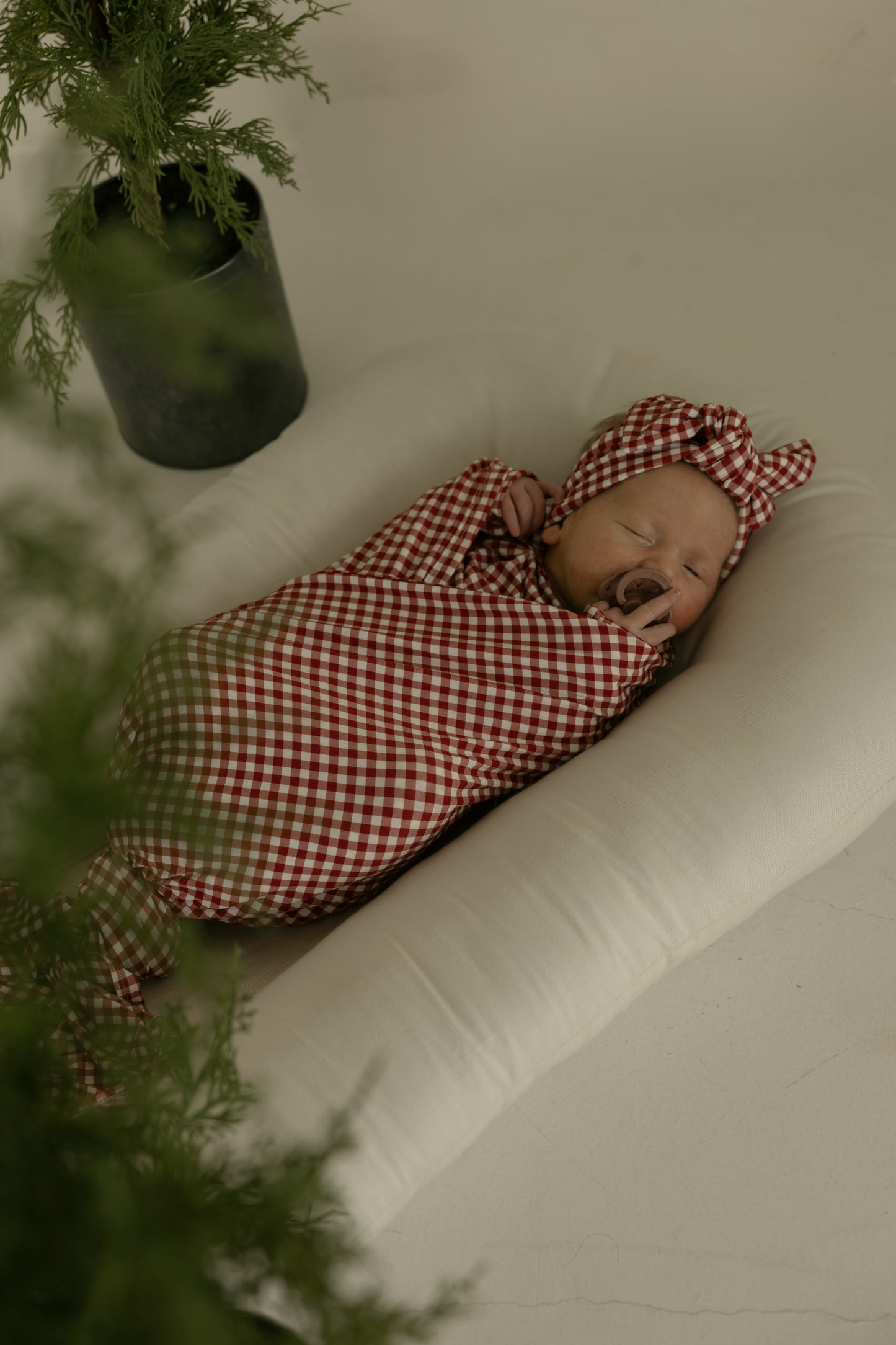 A sleeping baby wrapped in the forever french baby Bamboo Swaddle | Red Gingham with a matching headband rests on a soft white cushion, while green potted plants are partially visible in the foreground and background.