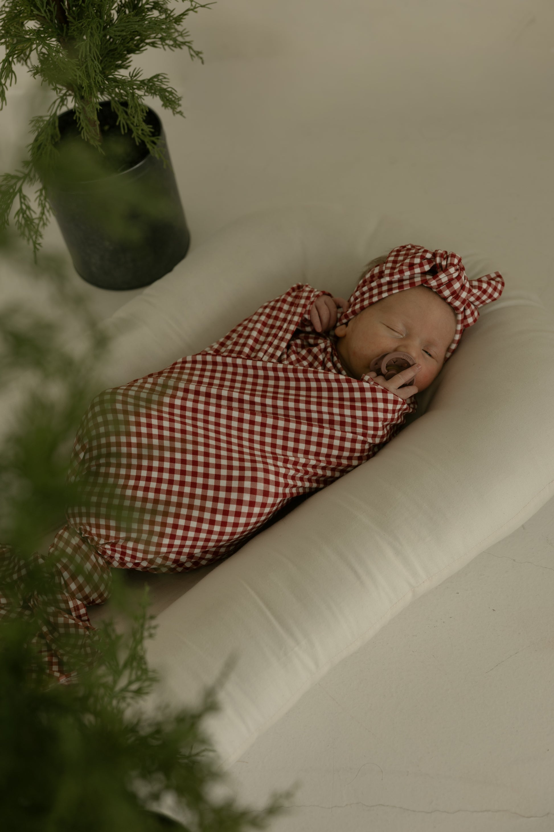 A sleeping baby wrapped in the forever french baby Bamboo Swaddle | Red Gingham with a matching headband rests on a soft white cushion, while green potted plants are partially visible in the foreground and background.