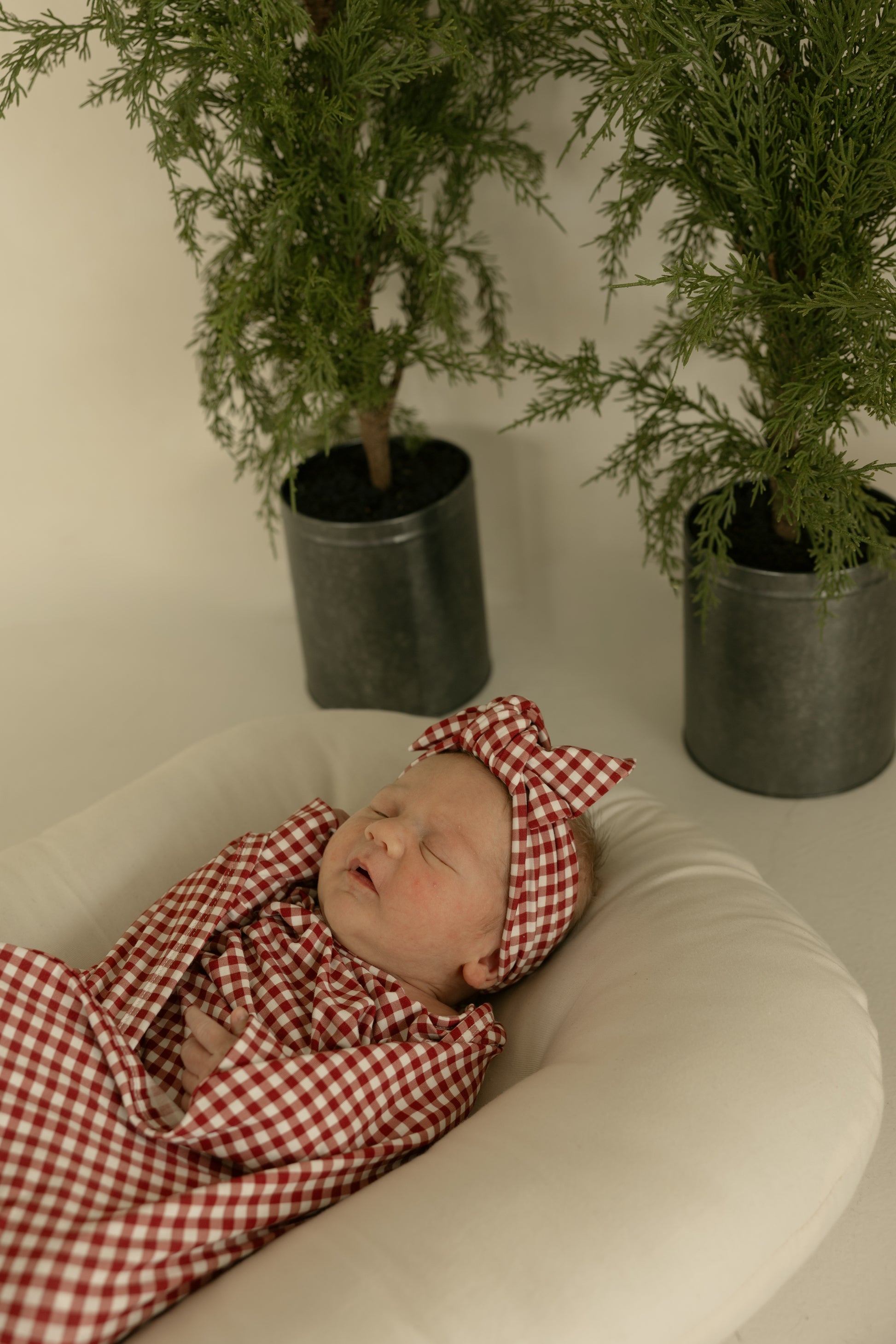 A baby wearing the forever french baby Bamboo Head Wrap in Red Gingham sleeps peacefully on a cushion, with two potted green plants in the background.