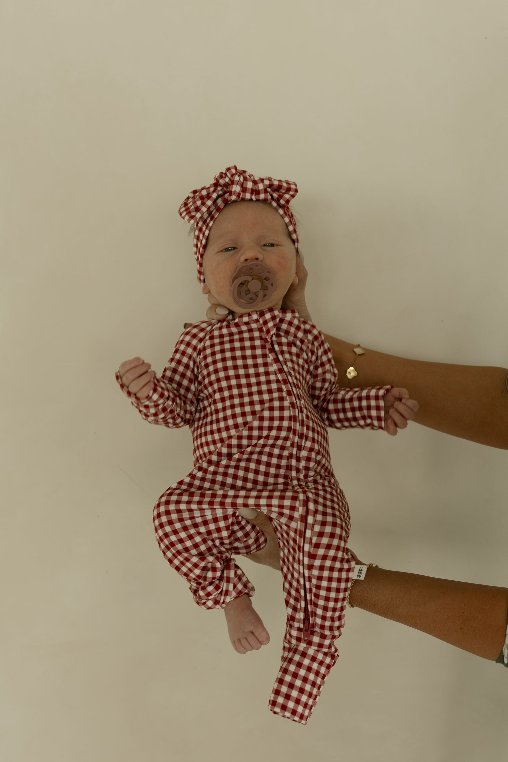 A baby lies on a light surface, wearing forever french baby’s Bamboo Zip Pajamas in Red Gingham with a matching headband. Supported by adult hands, the baby uses a pacifier.