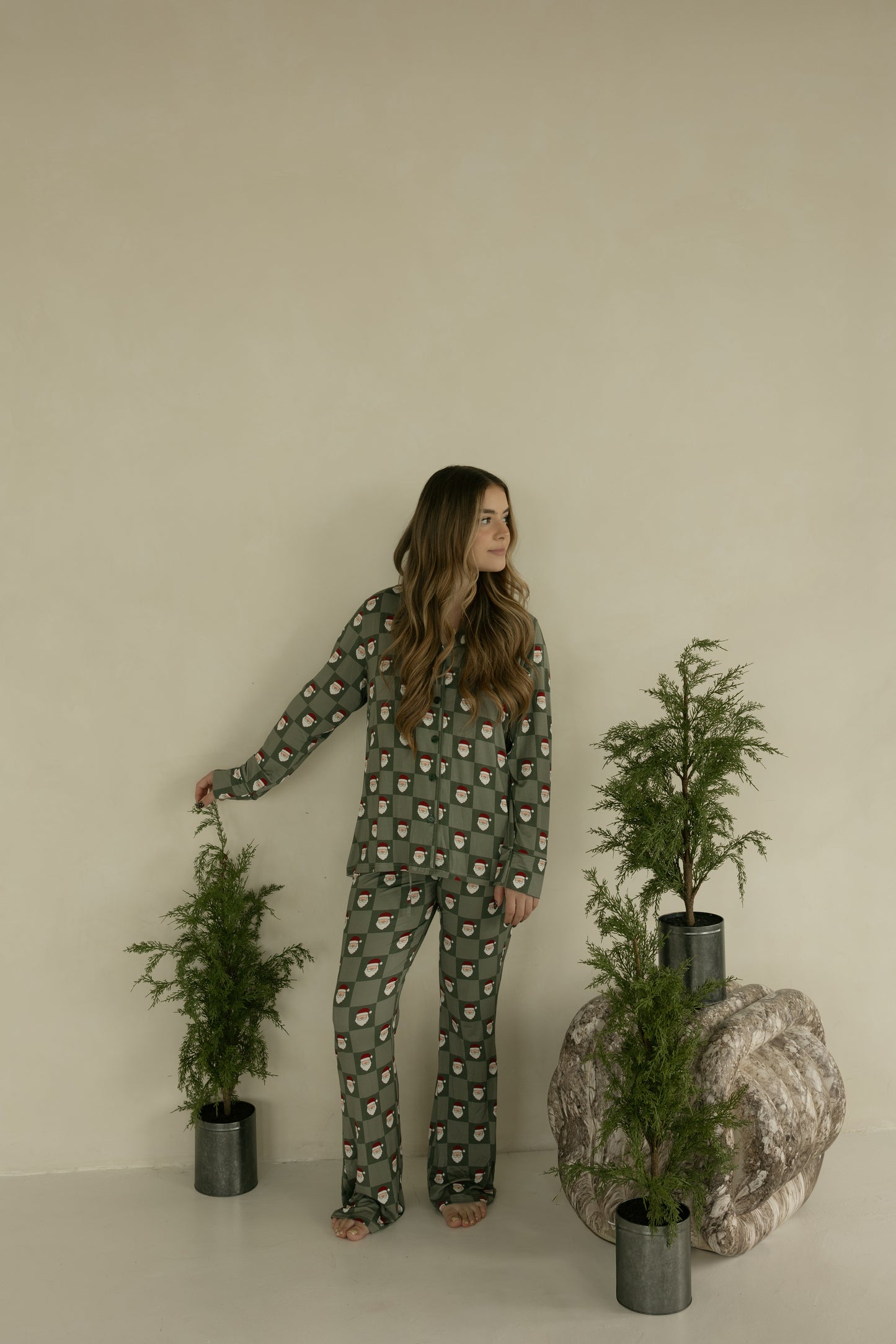 A woman in forever french baby’s Long Sleeve Women's Bamboo Pajamas | Santa Checkerboard stands indoors by a potted plant, surrounded by three lush green plants against a beige wall.