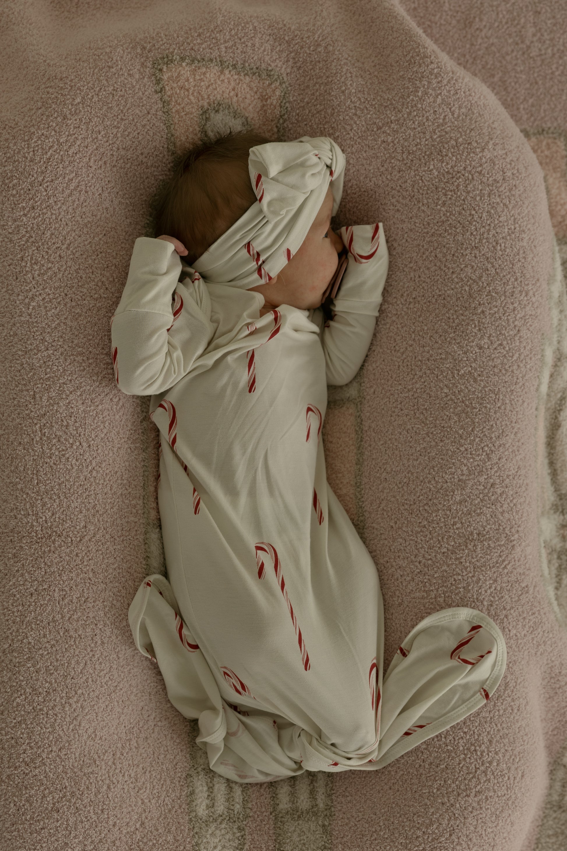 A baby in the forever french baby Bamboo Knotted Gown | Candy Cane, featuring red candy cane prints, lies on a soft pink blanket with one arm partly covering their face and their hat pulled over their forehead.