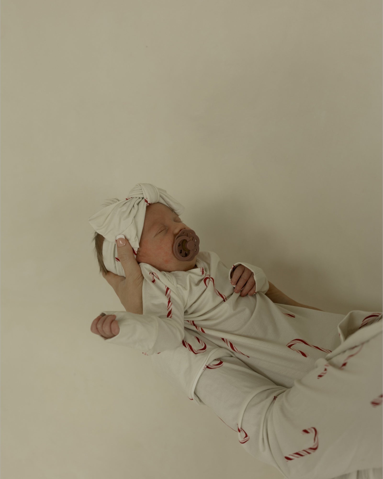 A newborn with a pacifier wears the Forever French Baby Bamboo Head Wrap | Candy Cane and matching white pajamas with red candy canes, cradled in someone's arms against a neutral background.