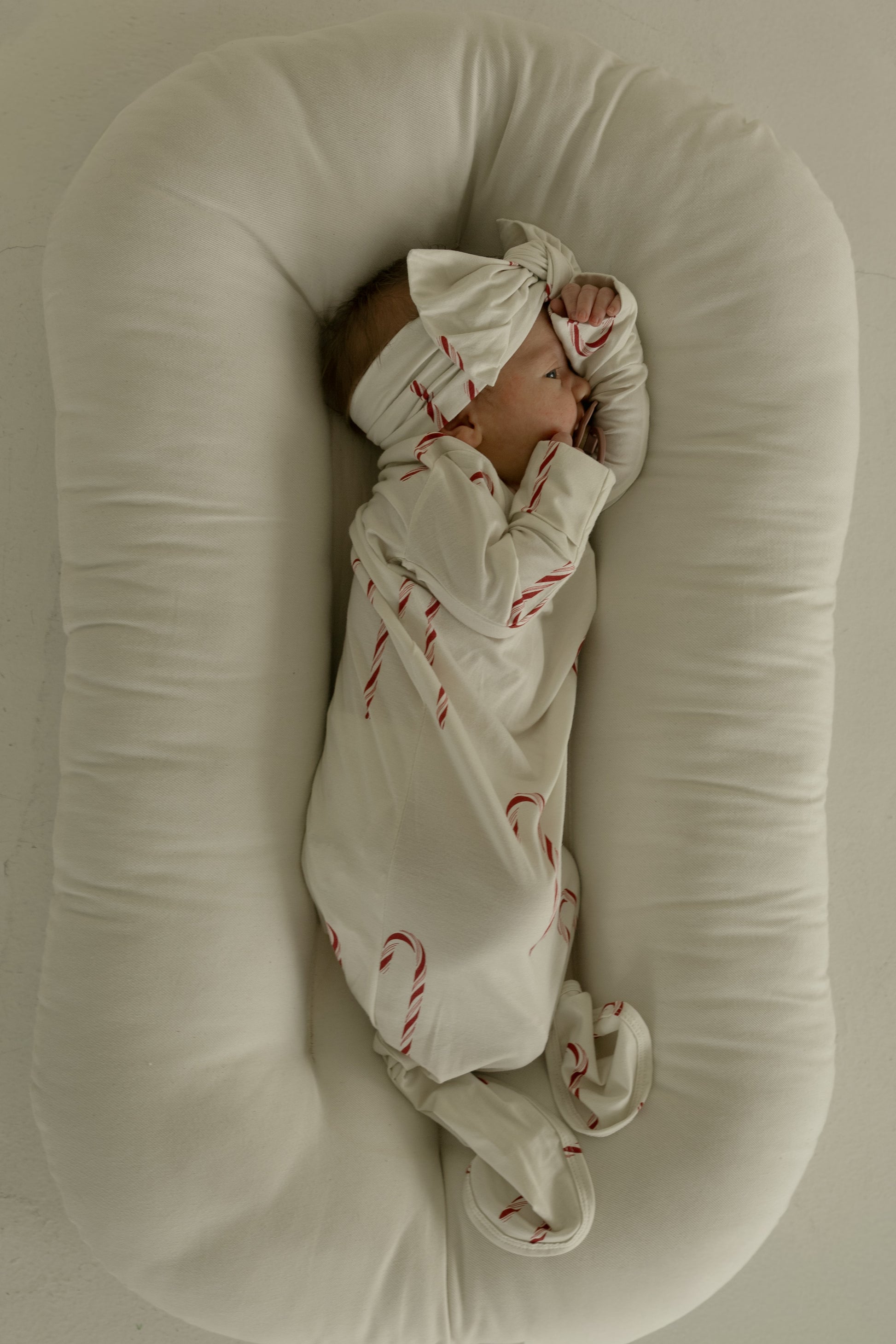 A baby in the forever french baby Bamboo Knotted Gown | Candy Cane—cream-colored with red candy cane patterns and a matching headband—lies sideways on a soft, white oval cushion with hands near their face.