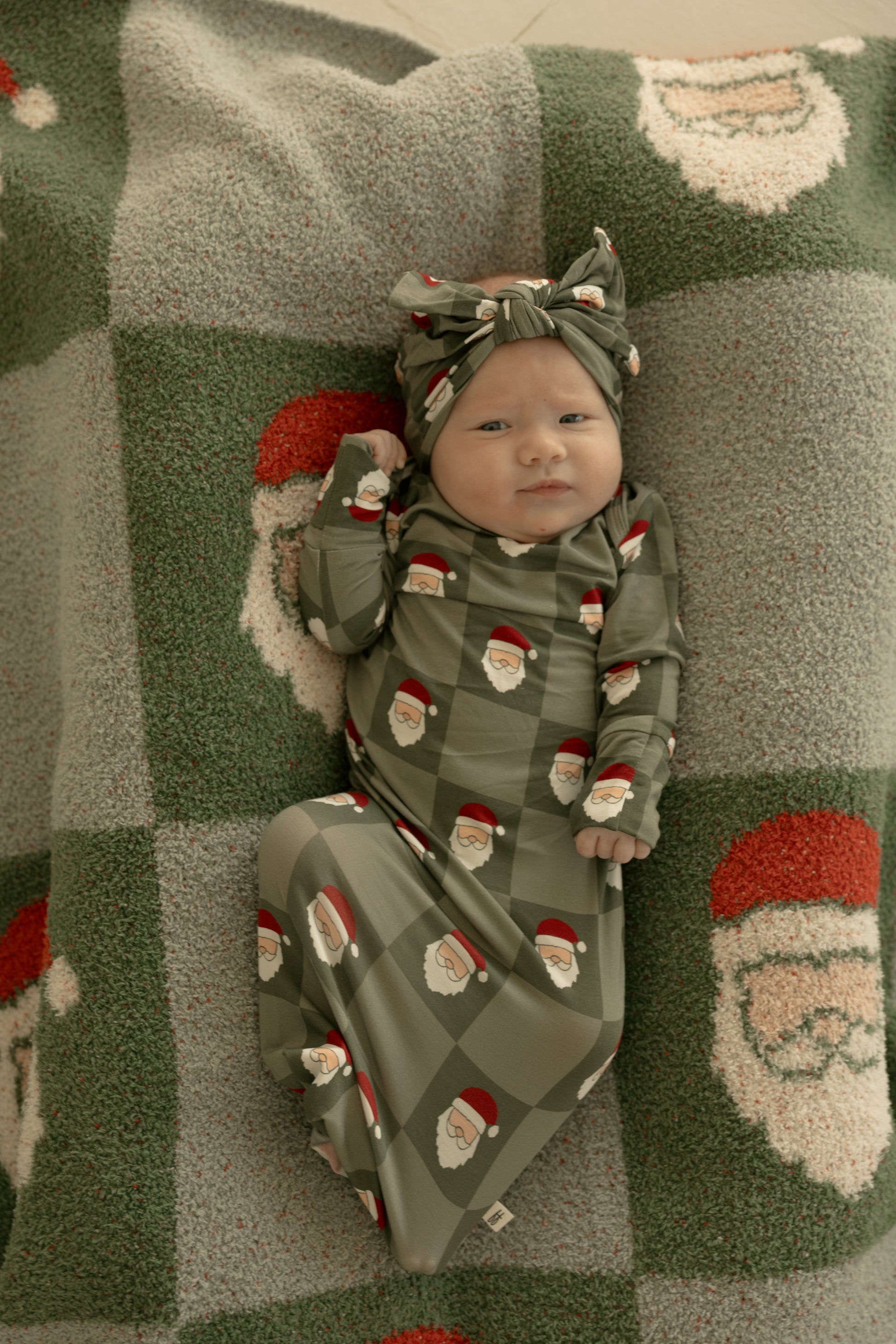 A newborn baby lies on a green and gray Santa-themed blanket, wearing a matching green outfit and the Bamboo Head Wrap | Santa Checkerboard by forever french baby, both adorned with festive Santa faces.