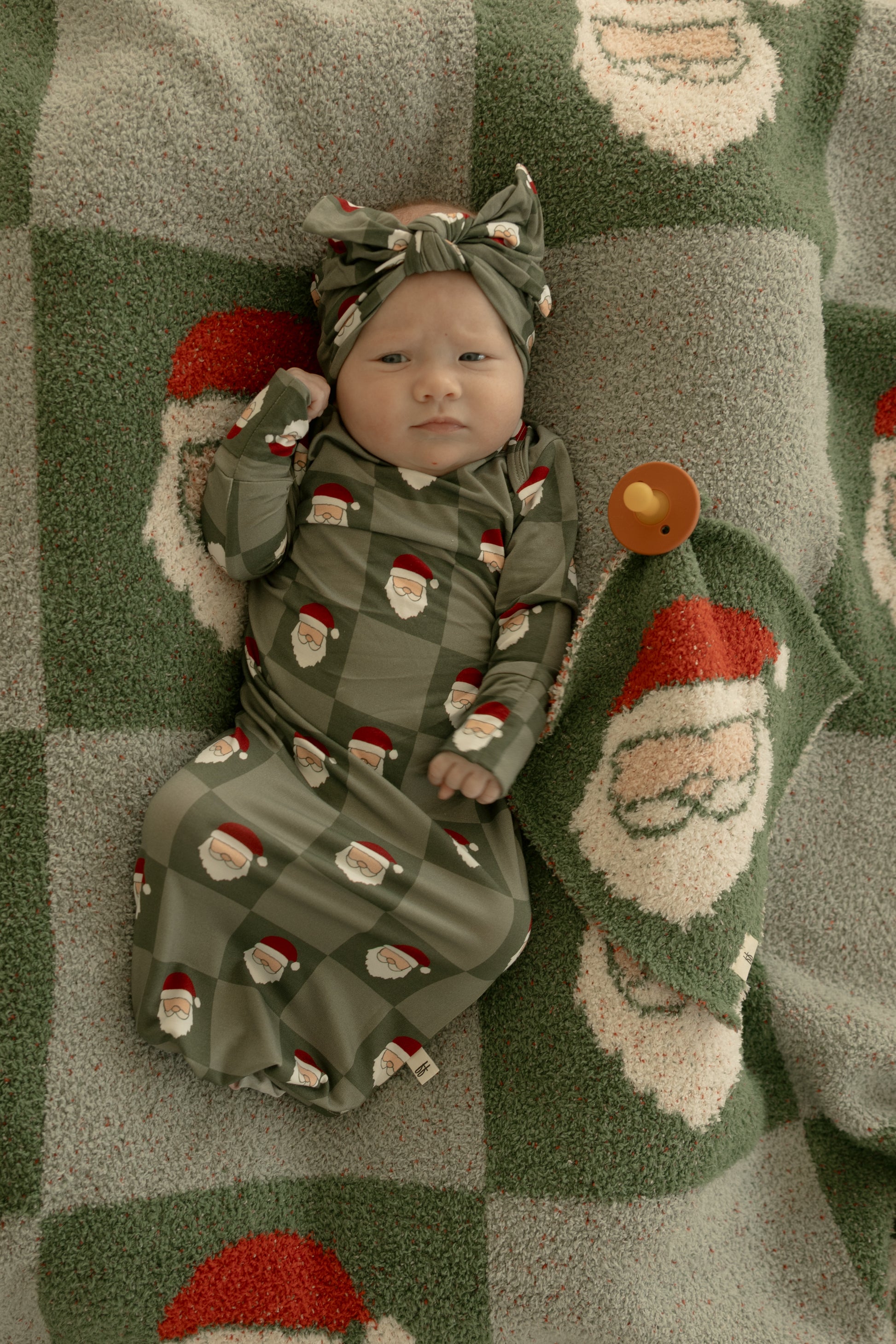A baby in a green forever french baby outfit and the Bamboo Head Wrap | Santa Checkerboard lies on a green and white blanket with large Santa faces. A brown pacifier is next to the baby.