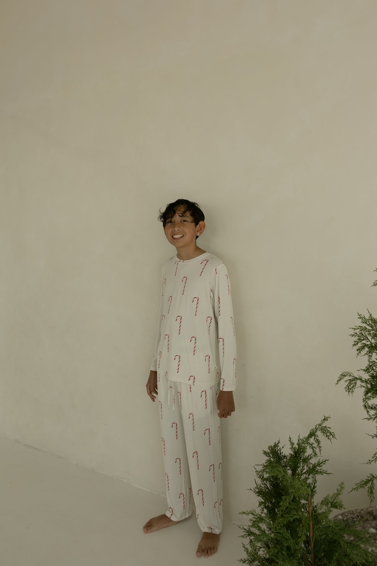 A boy wears forever french baby Pre-Teen Straight Leg Bamboo Pajamas in the Candy Cane pattern, standing barefoot indoors by a green plant and smiling against a plain beige wall.