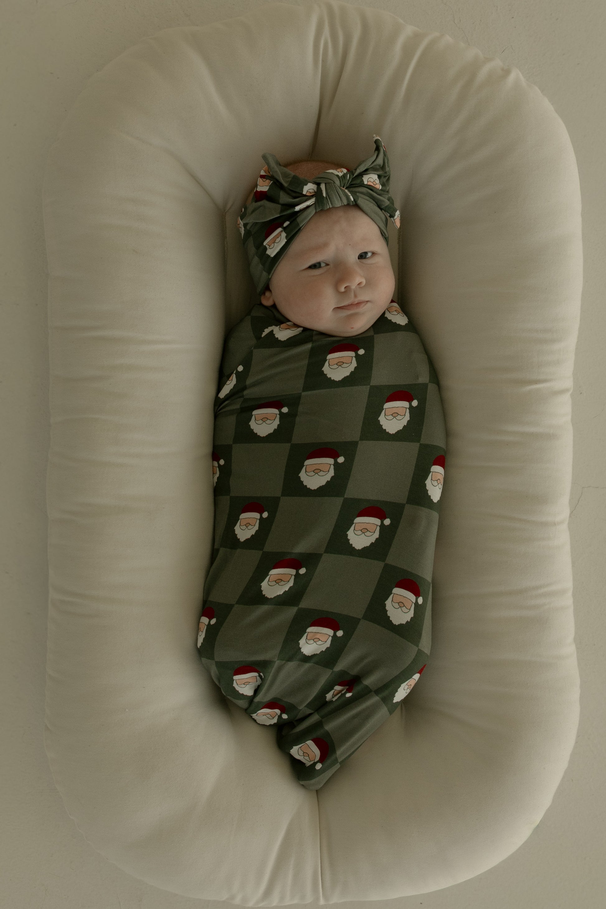 A baby in the forever french baby Bamboo Swaddle | Santa Checkerboard, with a matching headband, rests on a soft oval white cushion against a light background.