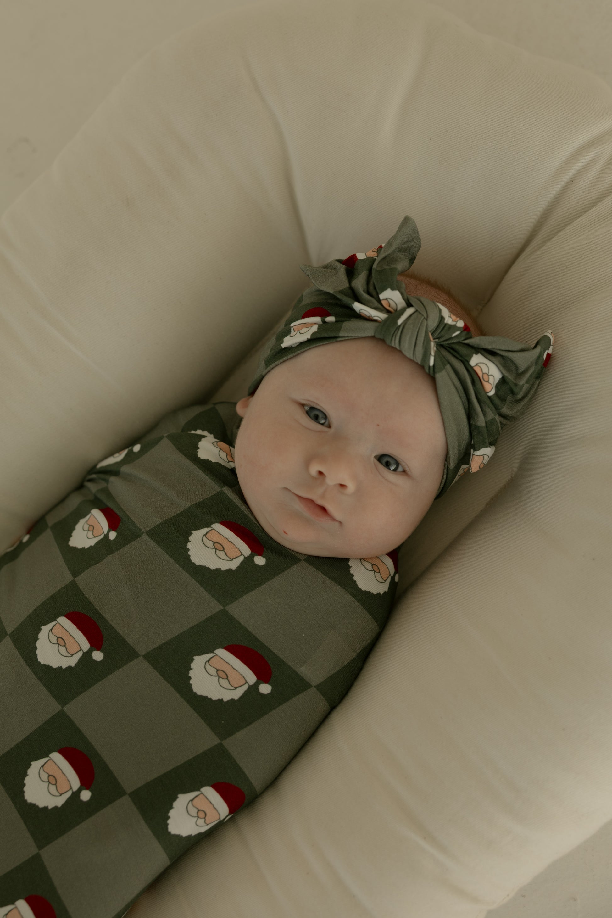 A baby, wrapped in the forever french baby Bamboo Swaddle | Santa Checkerboard and matching headband, lies on a soft cream cushion.