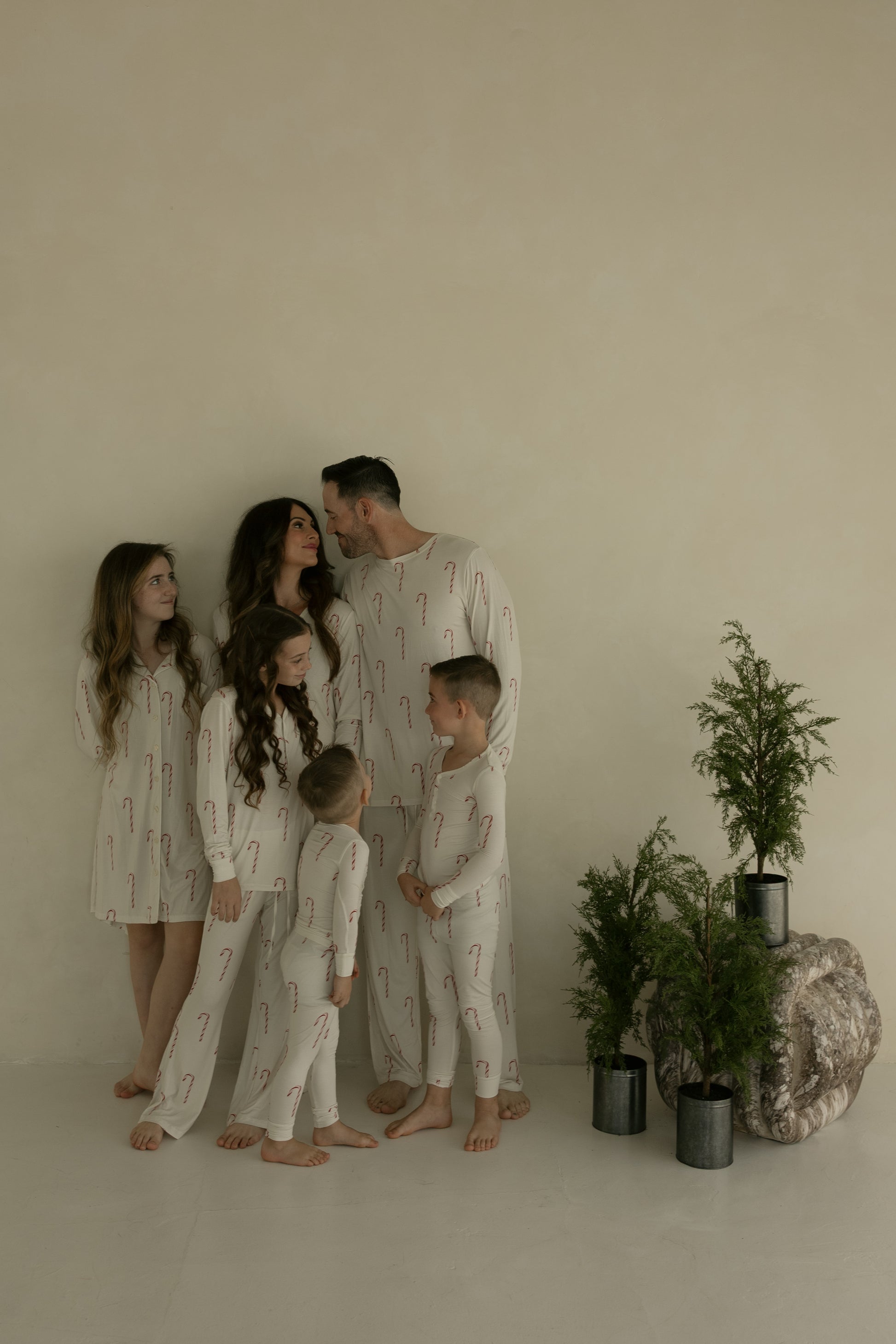 A family of six, barefoot and joyful, poses by potted evergreens in matching forever french baby Pre-Teen Flare Bamboo Pajamas | Candy Cane, featuring red candy canes, against a plain beige wall.