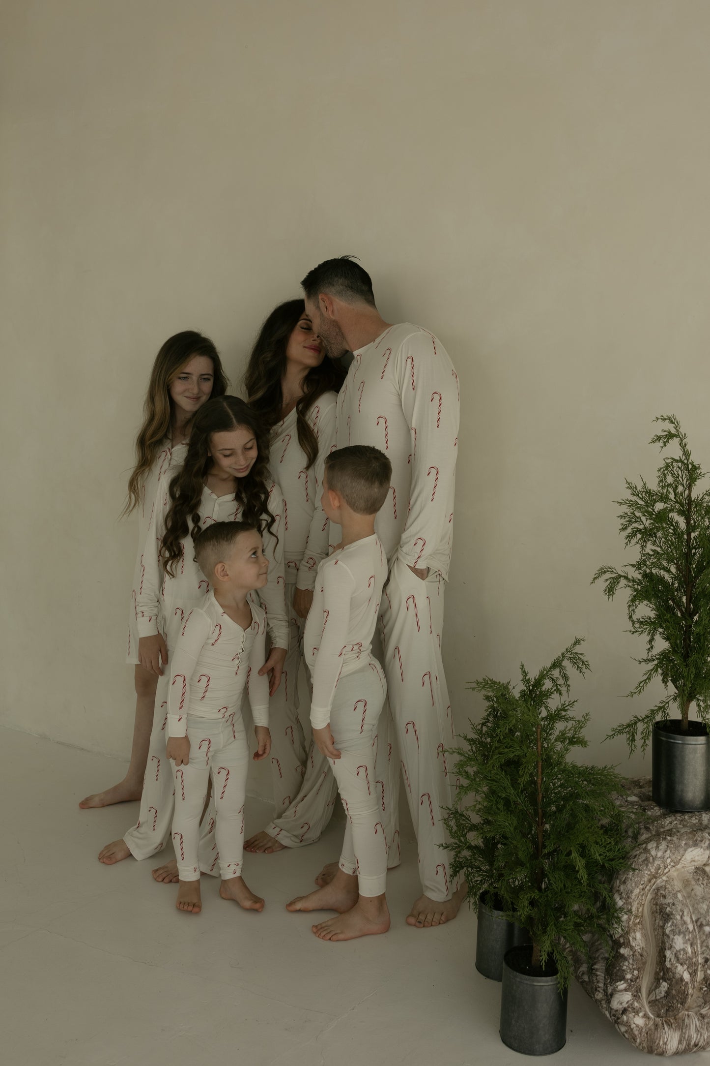 A family of six wears forever french baby's Bamboo Two Piece Pajamas | Candy Cane, all matching in cream with red candy cane prints, as they stand barefoot together by potted evergreens while the parents share a kiss and the kids smile.