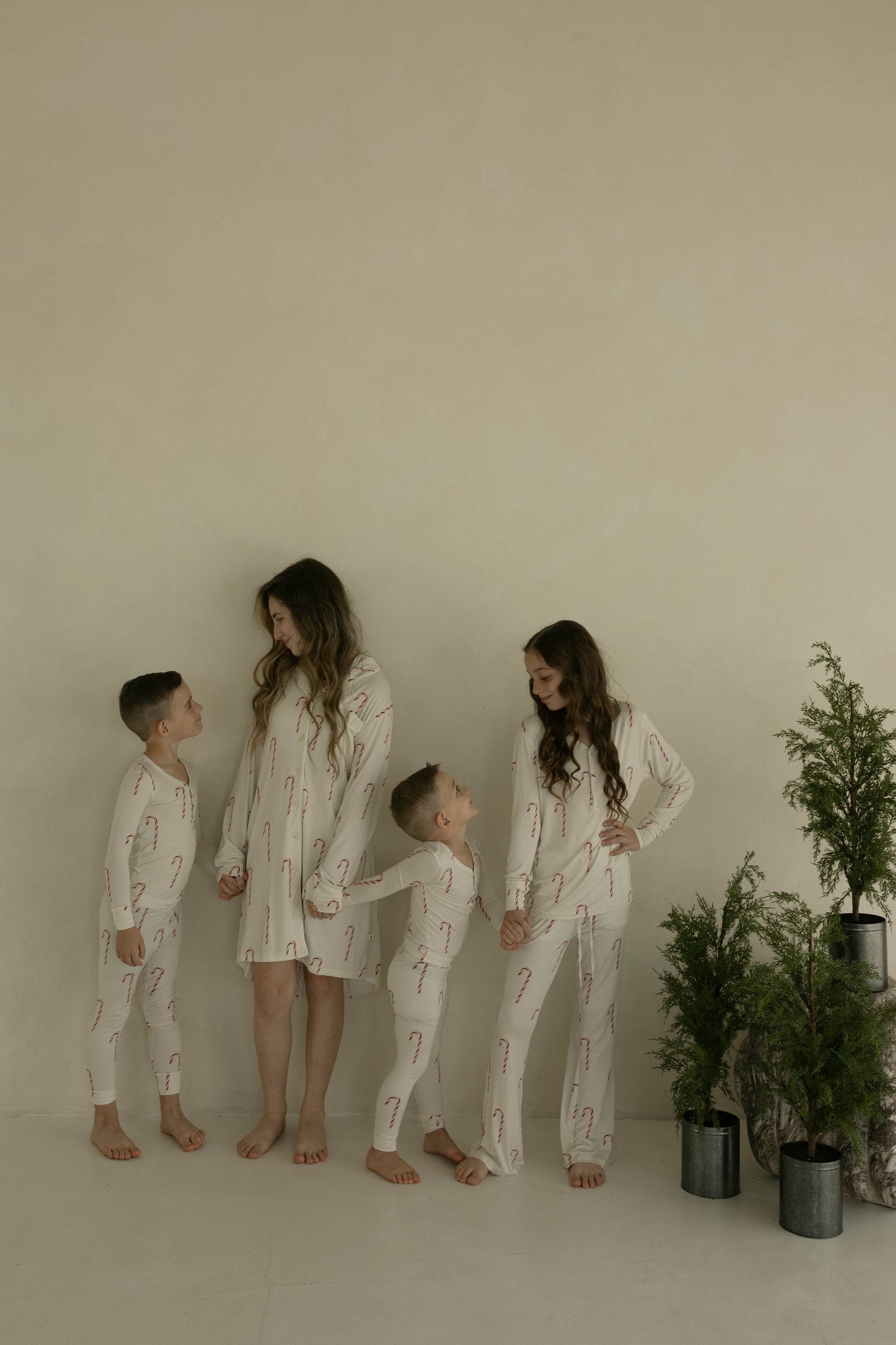 Four children smile barefoot against a beige wall, holding hands in matching forever french baby Pre-Teen Flare Bamboo Pajamas | Candy Cane, featuring cream fabric with red candy canes. Small potted evergreens sit to the right.