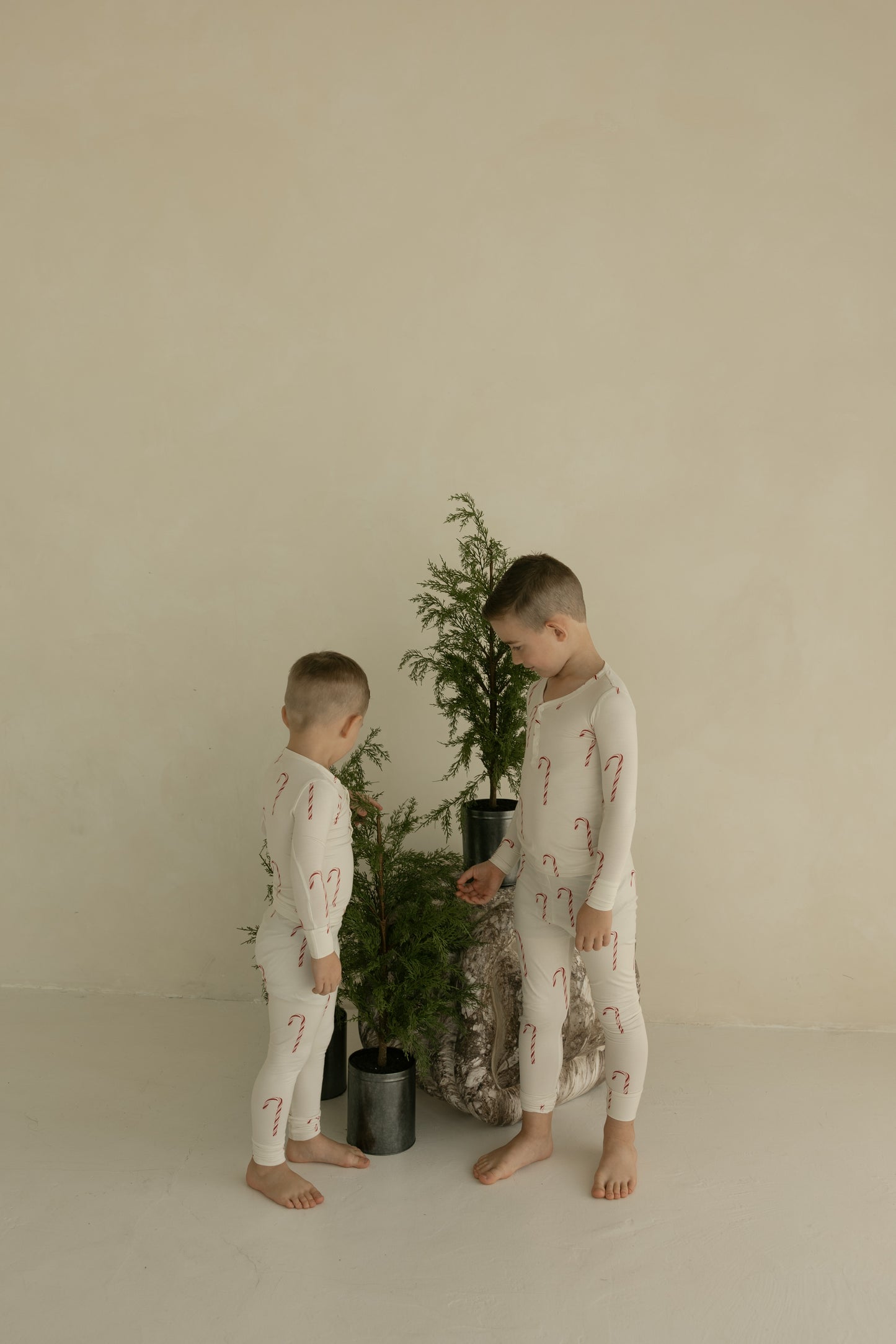 Two children in matching forever french baby Bamboo Two Piece Pajamas | Candy Cane stand barefoot by potted evergreens, set against a simple beige backdrop—ideal holiday sleepwear for cozy nights.