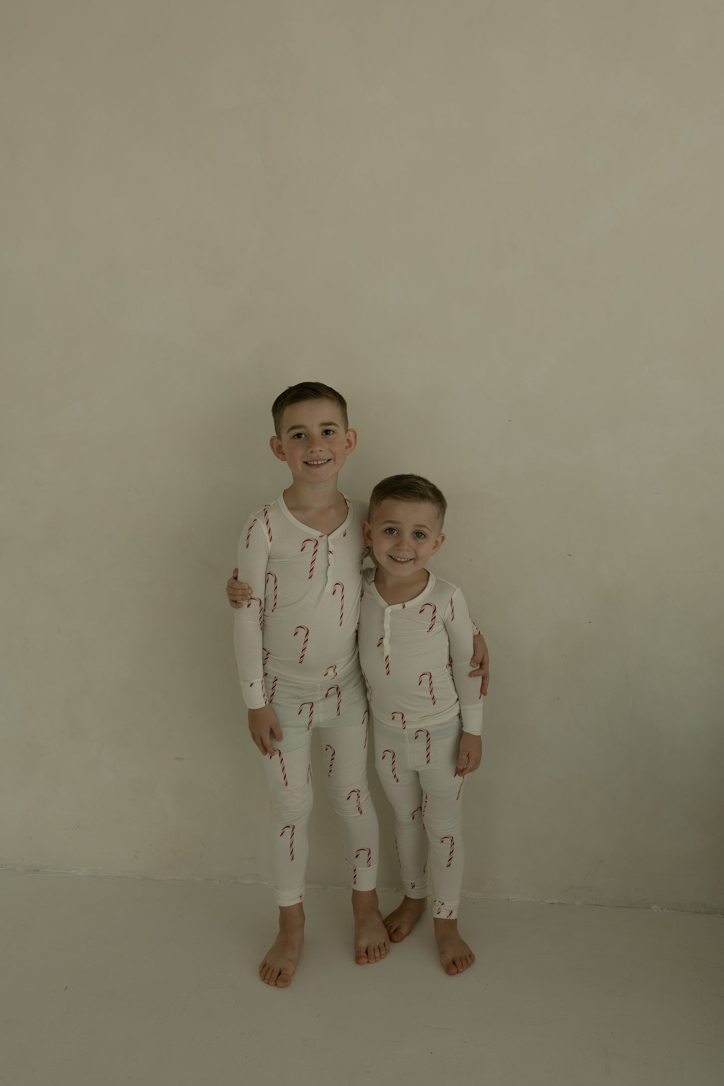 Two kids smile at the camera in matching forever french baby Bamboo Two Piece Pajamas | Candy Cane—cream-colored, hypo-allergenic bamboo sleepwear perfect for children, shown barefoot on a light beige background.