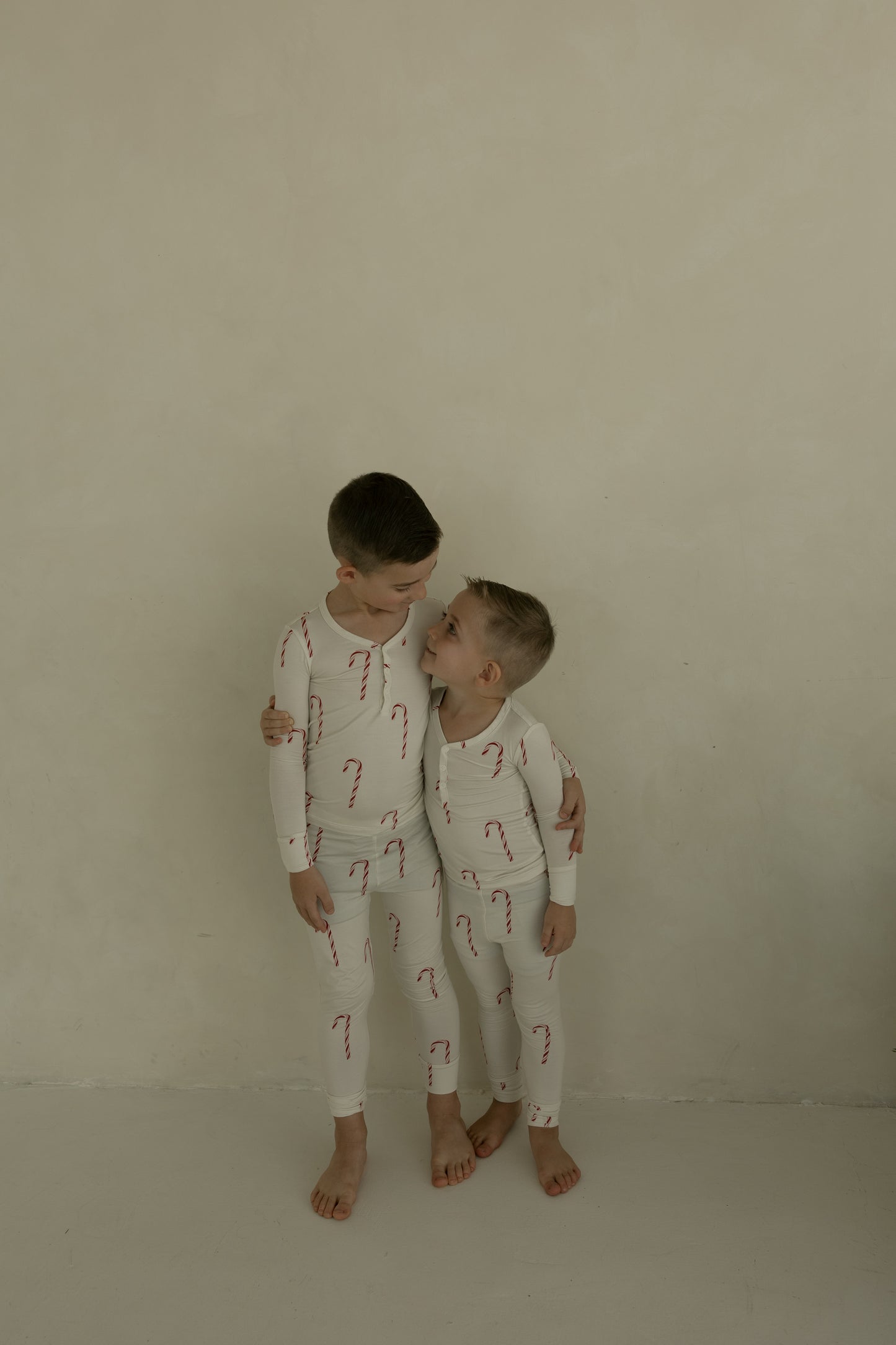 Two young boys, barefoot against a plain wall, wear matching forever french baby Bamboo Two Piece Pajamas | Candy Cane. The older boy hugs the younger as they smile at each other in festive, cozy kids sleepwear.