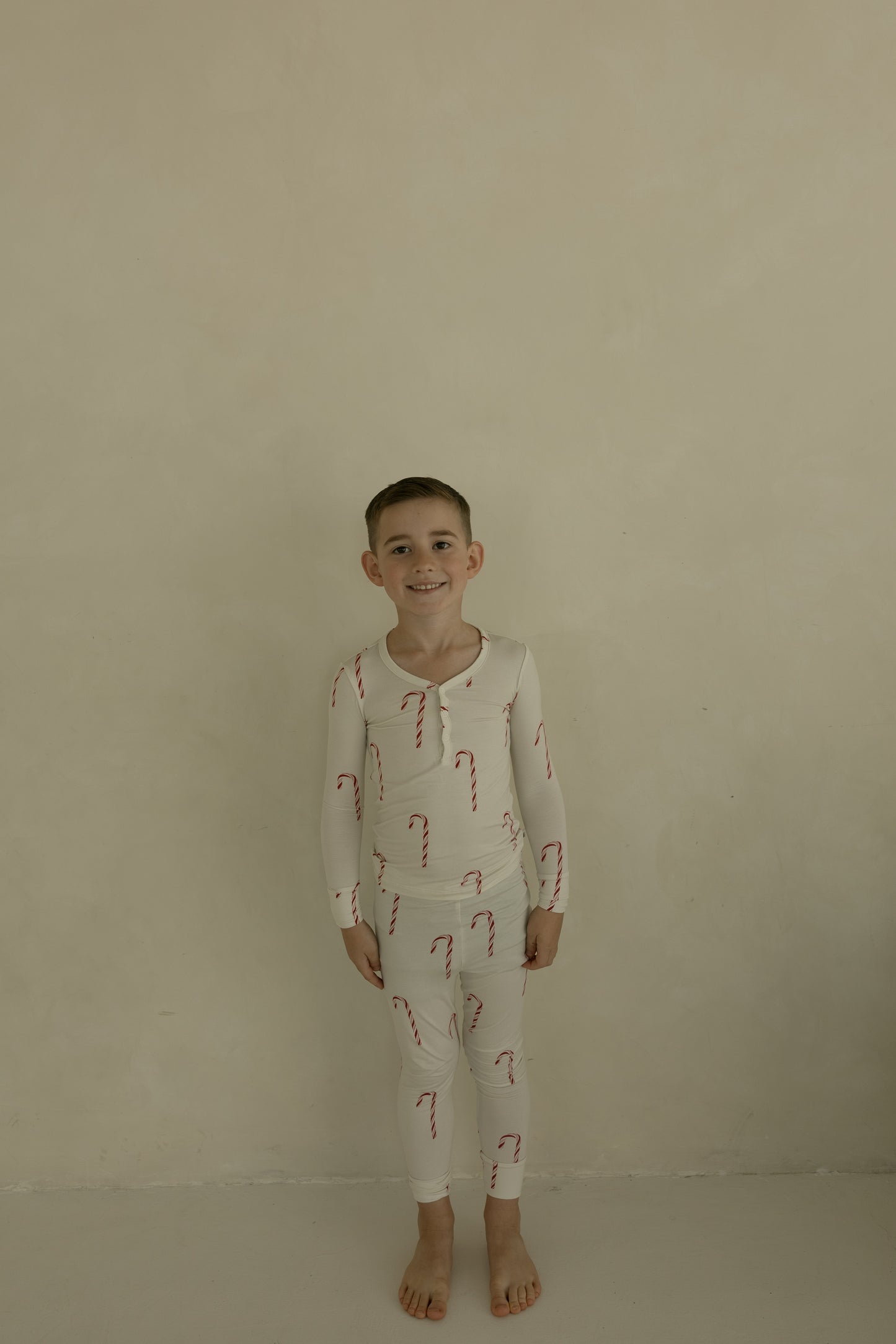 A young child smiles barefoot against a beige wall, wearing forever french baby’s Bamboo Two Piece Pajamas | Candy Cane—soft, festive kids sleepwear that’s gentle on skin.