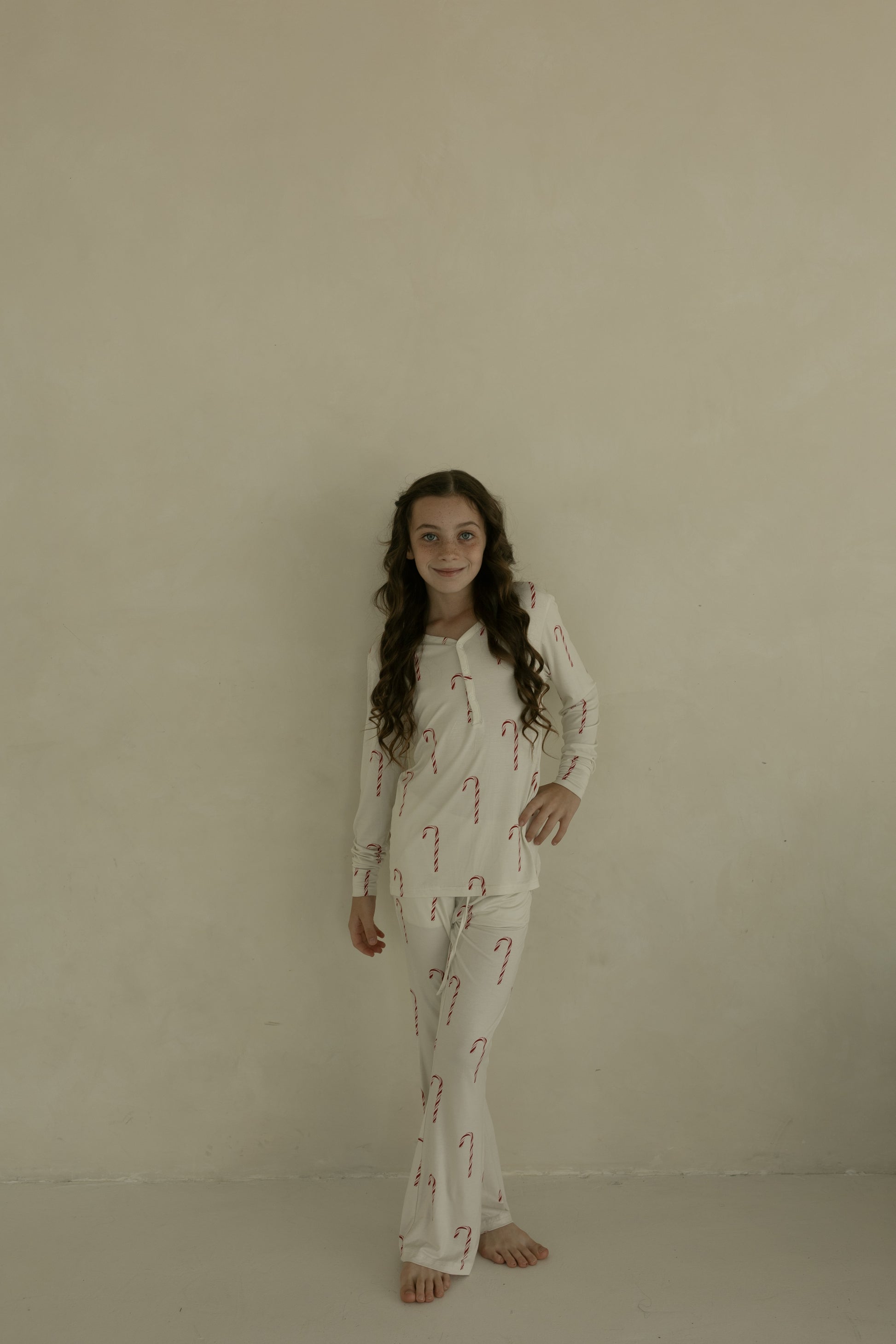 A young girl with long curly hair stands barefoot by a cream wall, smiling gently in forever french baby Pre-Teen Flare Bamboo Pajamas | Candy Cane.