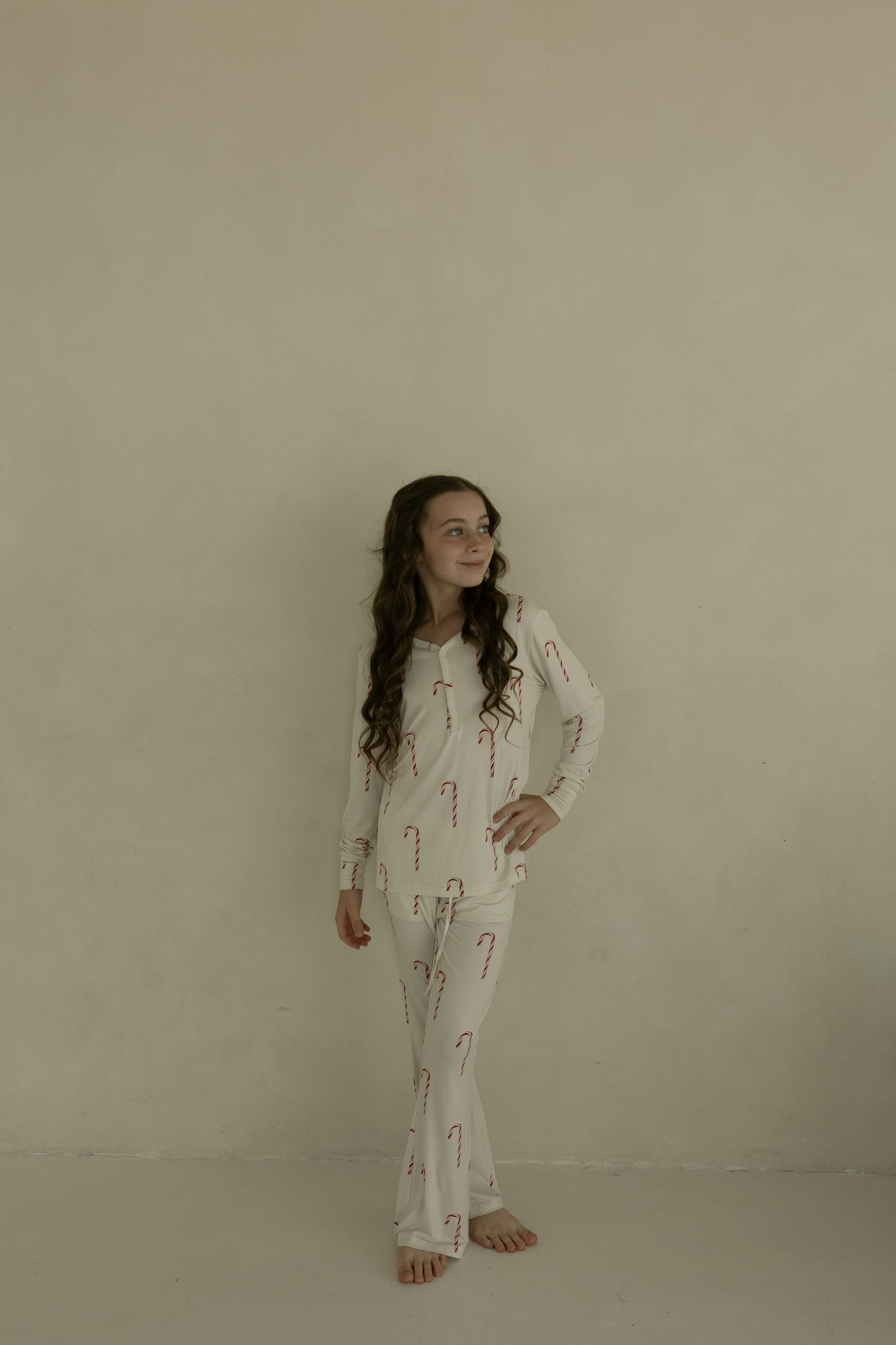 A young girl with long wavy hair stands barefoot by a beige wall, wearing forever french baby’s Pre-Teen Flare Bamboo Pajamas in the Candy Cane print—cozy, hypoallergenic sleepwear perfect for the holidays.