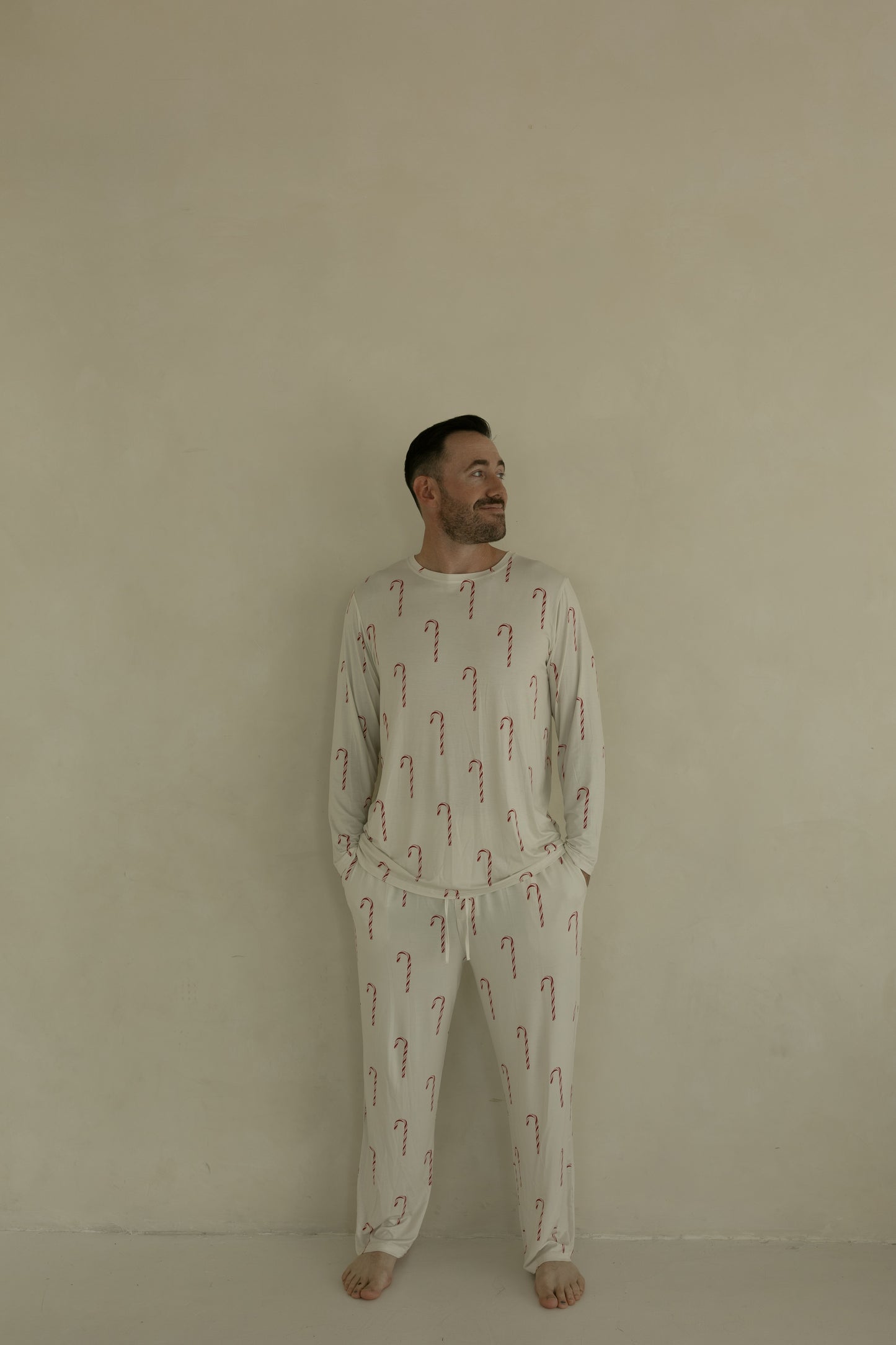 A man stands barefoot against a plain beige wall, wearing forever french baby Men's Bamboo Pajamas | Candy Cane, featuring a light-colored fabric with red candy cane pattern. He has his hands in his pockets and is smiling slightly while looking to the side.